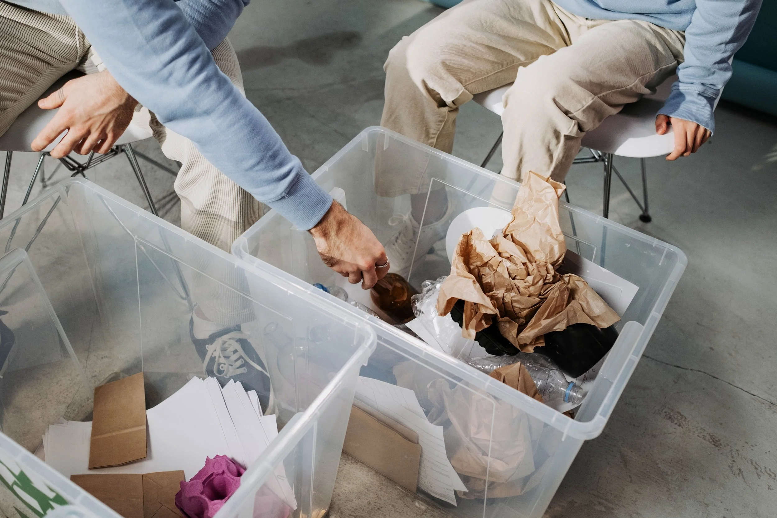 Two people sitting on chairs sorting items in plastic storage bins filled with papers, crumpled brown paper, and other assorted objects. One person is wearing a blue long-sleeve shirt and khaki pants, the other's clothing is partially visible.