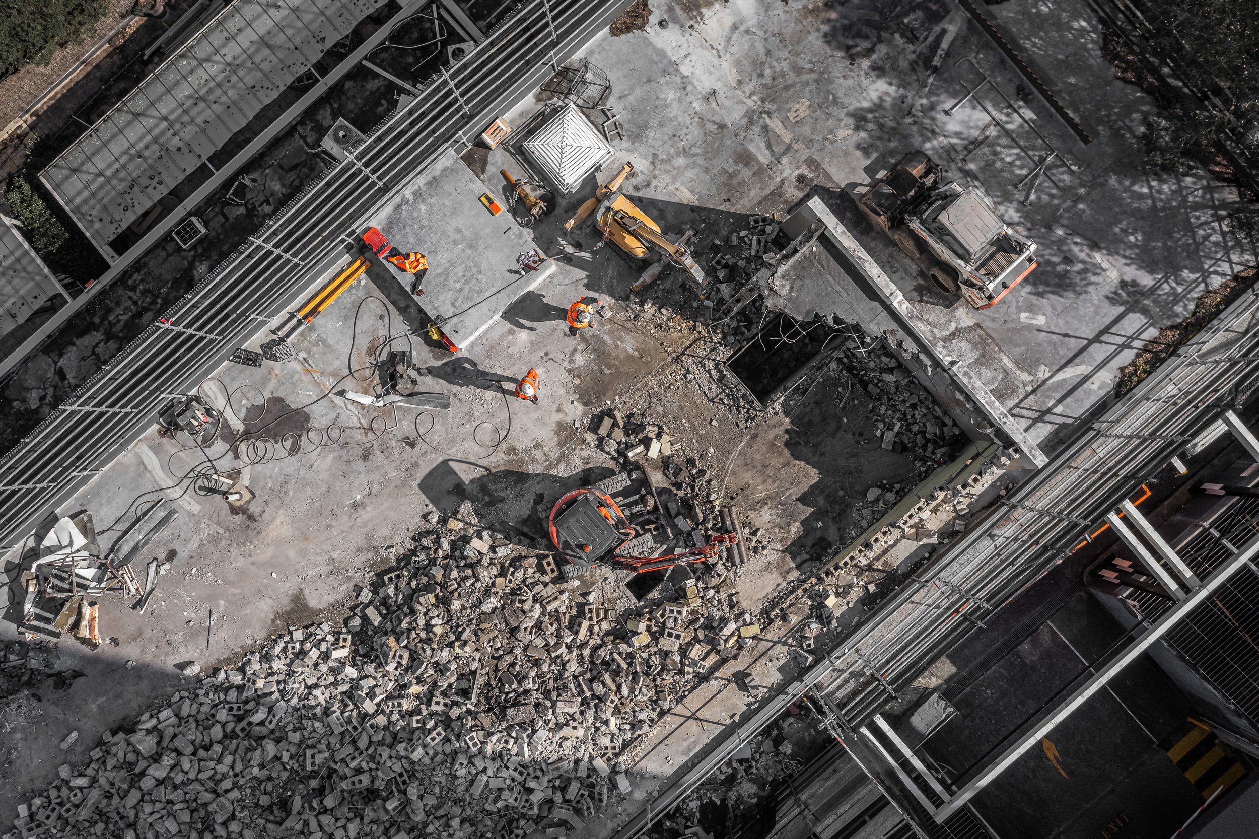 An aerial view of a construction site with workers in orange safety vests, machinery, and piles of broken concrete and bricks.