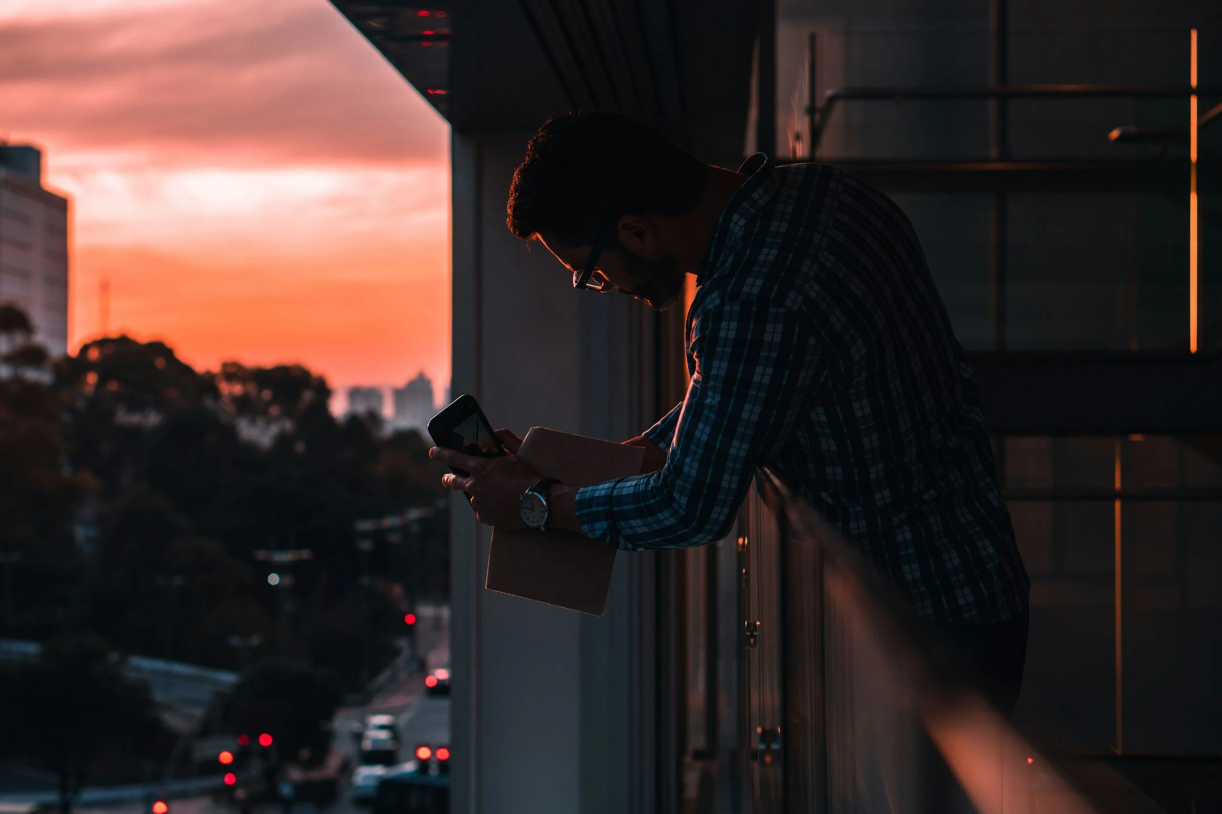A man wearing glasses and a checkered shirt is standing outside on a balcony or rooftop at sunset, looking at his phone, with city buildings and a busy street below.