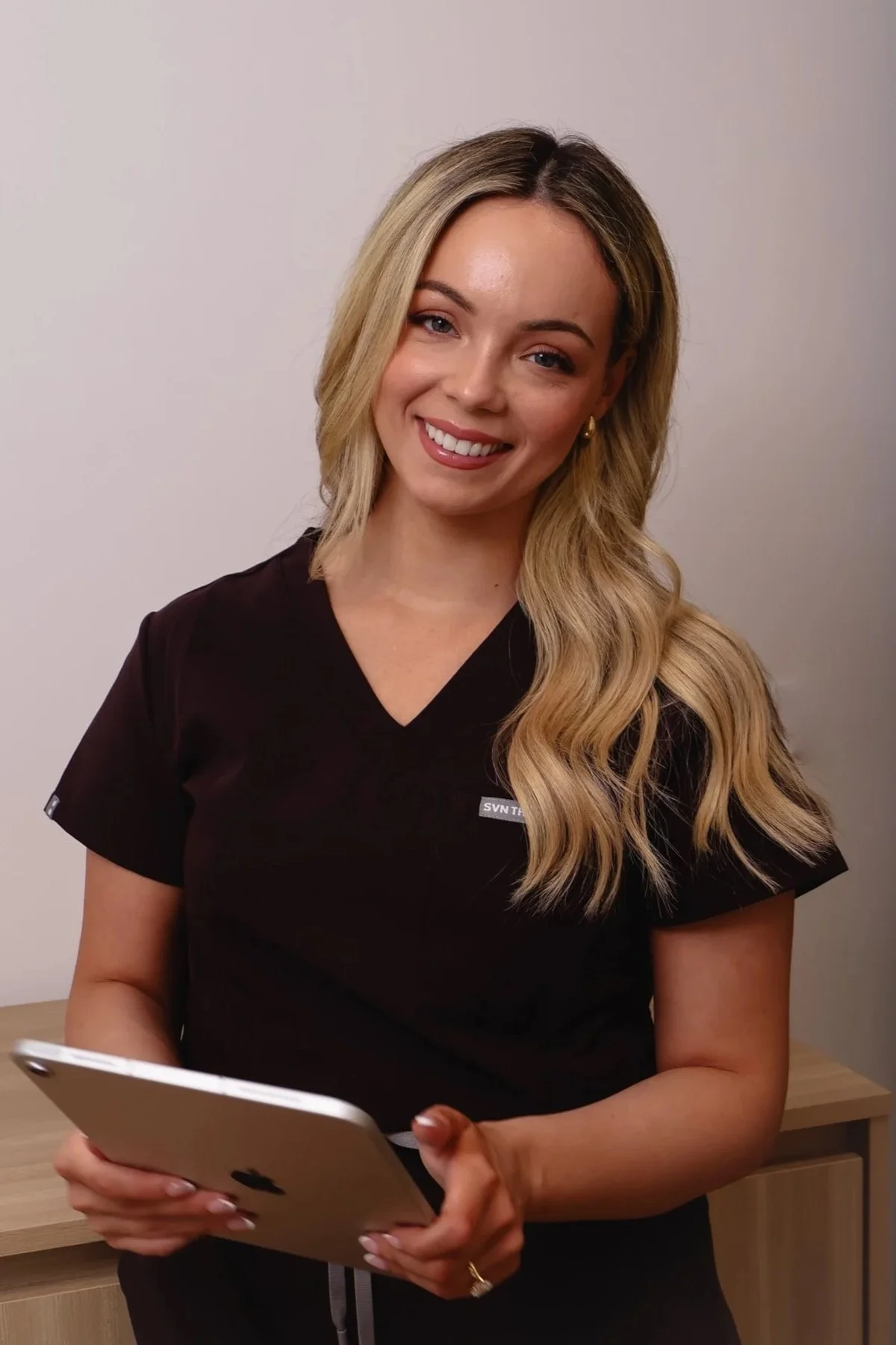 A woman with long blonde hair, wearing a black scrub top, holding a tablet device, standing indoors against a plain wall.