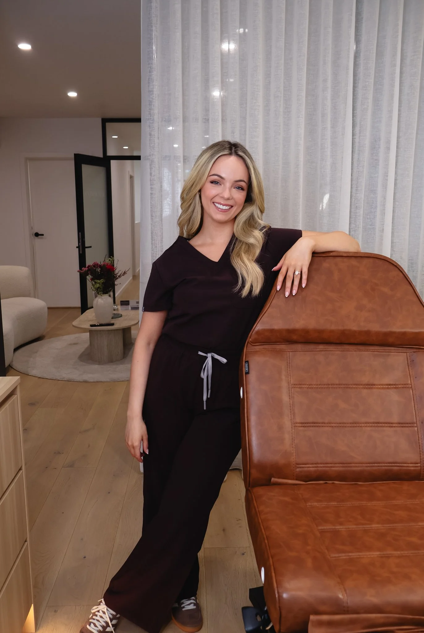 A woman with long blonde wavy hair, wearing a black scrubs top, black pants, and white sneakers, is leaning on a brown leather chair with her right arm, smiling and looking at the camera inside a modern living space with white curtains, a round beige rug, and a small wooden table with a vase of flowers.