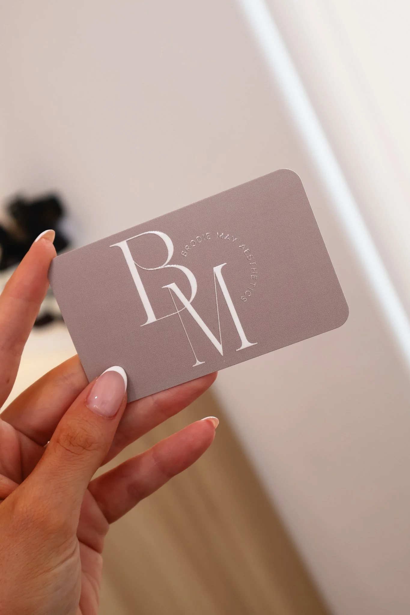 Person holding a gray card with white text and logo, showing the words "BODY ME ALLES" and stylized letters 'B', 'M', and 'V'.