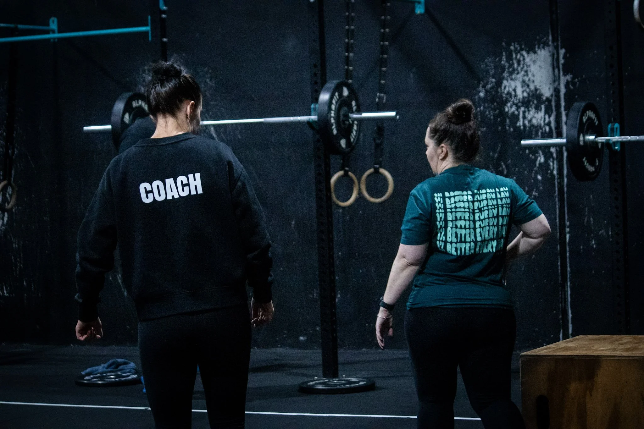 Two women standing in a gym near weightlifting equipment, one wearing a shirt labeled 'COACH' and the other in a teal shirt, with gym rings and a weightlifting bar in the background.