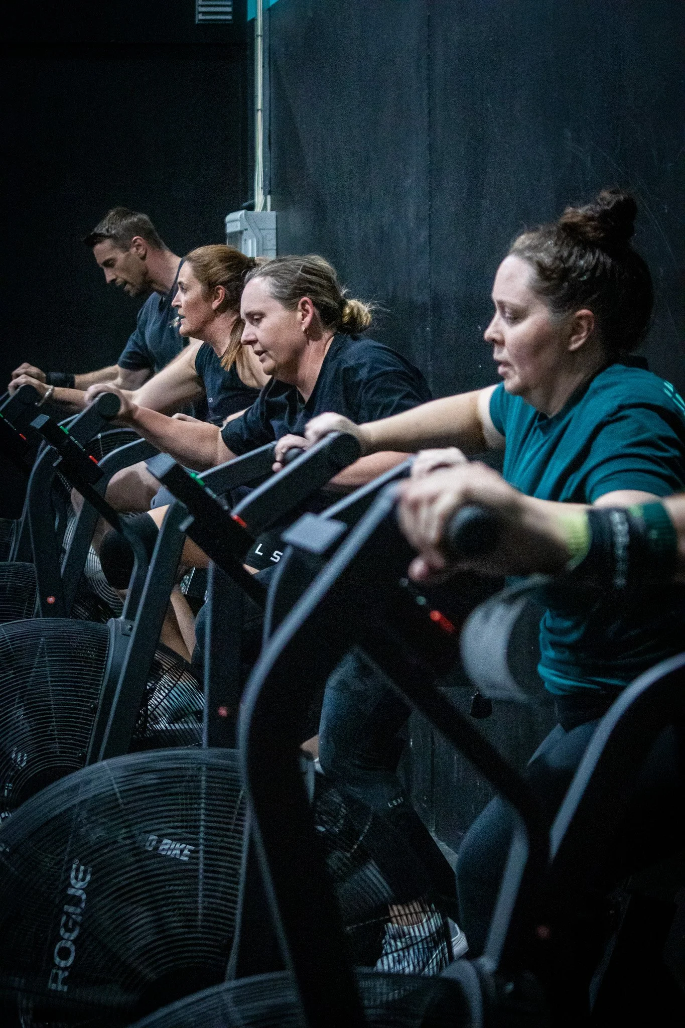 Four people are exercising on stationary bikes in a dark indoor gym. They are focused and pedaling, with a black wall behind them.