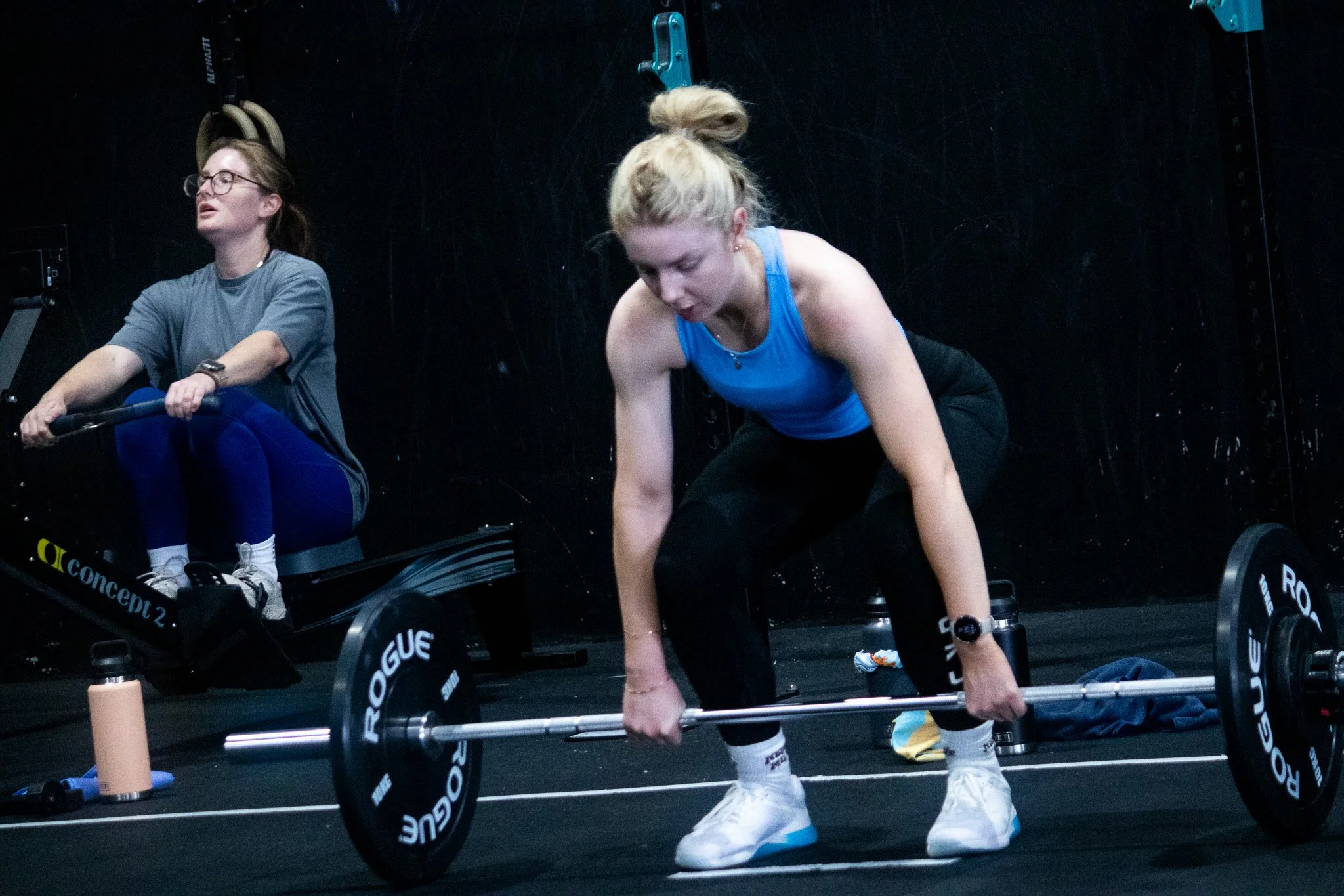 Two women engaging in a workout session at a gym, one is sitting on a rowing machine, and the other is preparing to lift a barbell, both are focused on their exercise routines.