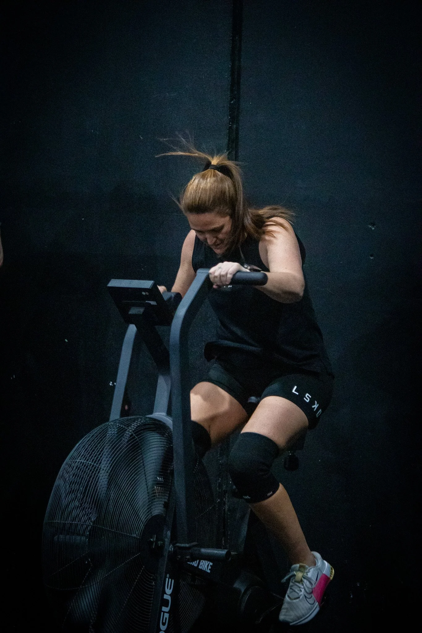 A woman in workout clothes is riding a stationary bike inside a gym, with black walls and a large fan in front of her.