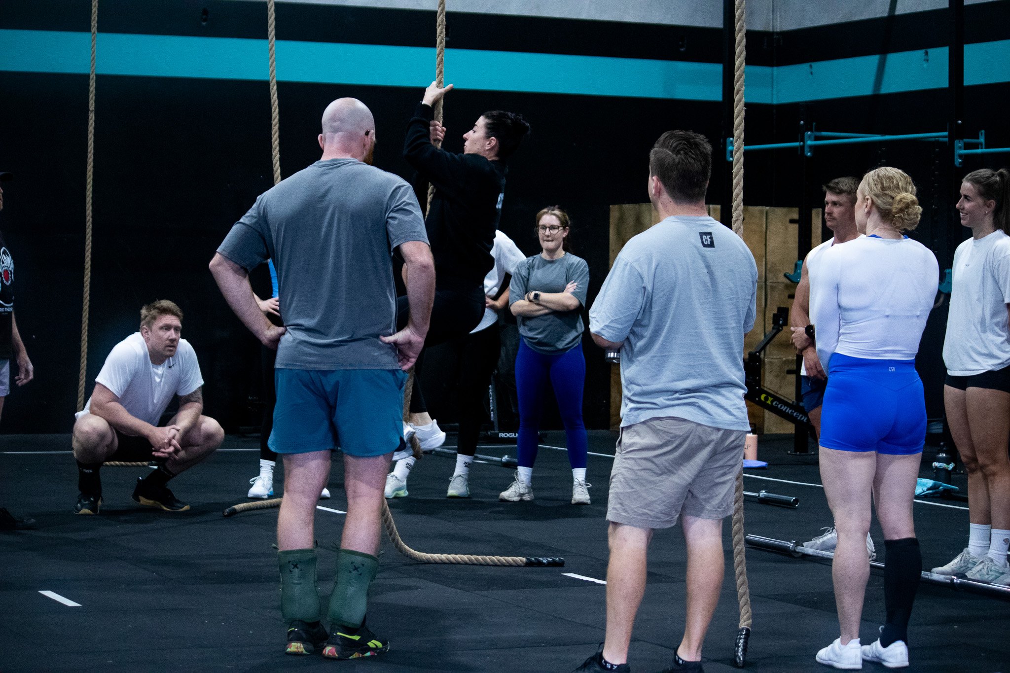 Group of people in a gym participating in a workout session, some using ropes for exercises, while others observe or rest.