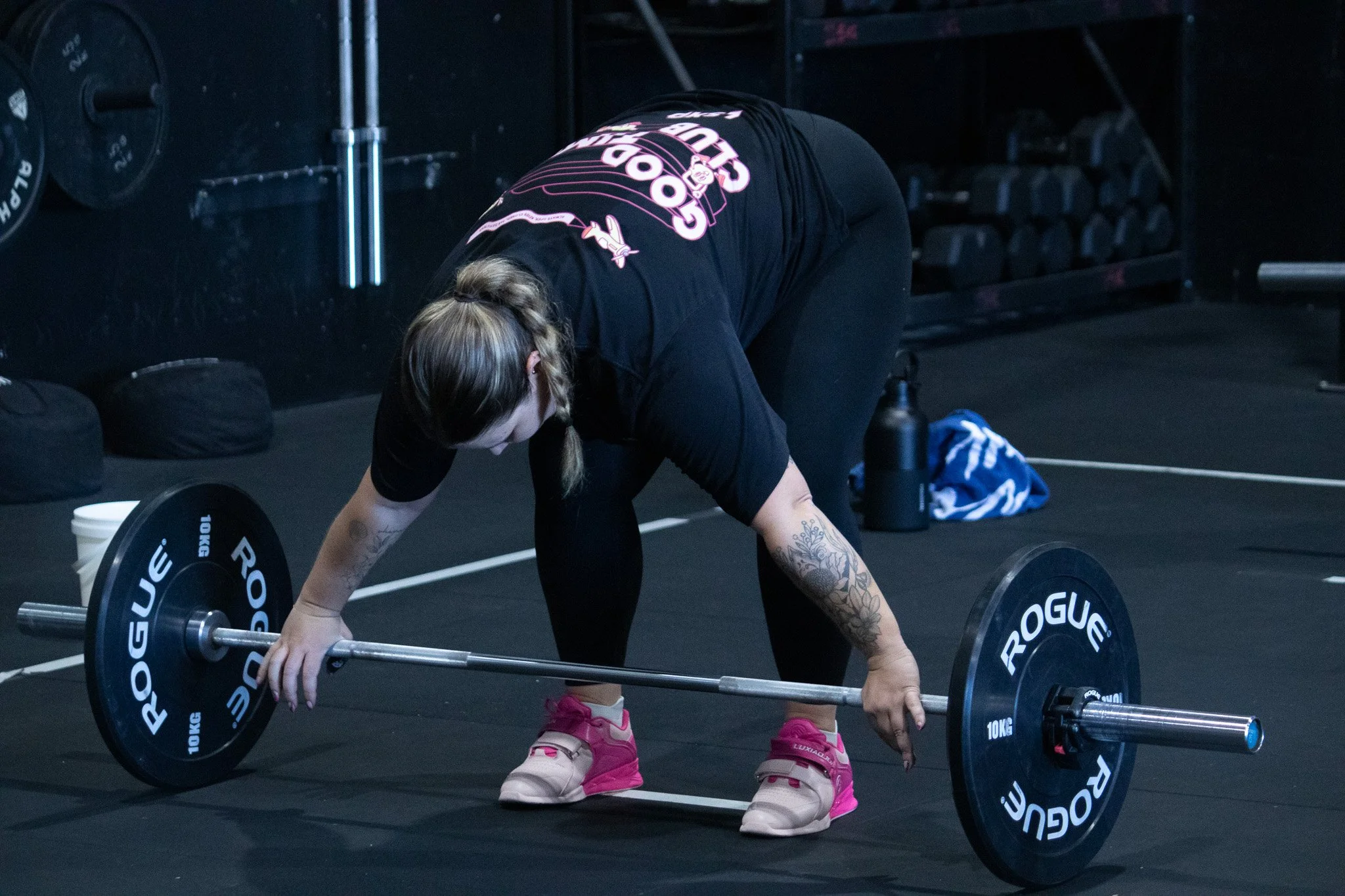 A woman with blonde hair in a ponytail, tattoos on her arm, wearing black athletic clothing and pink sneakers, is bending over and lifting a barbell with black weight plates in a gym setting.
