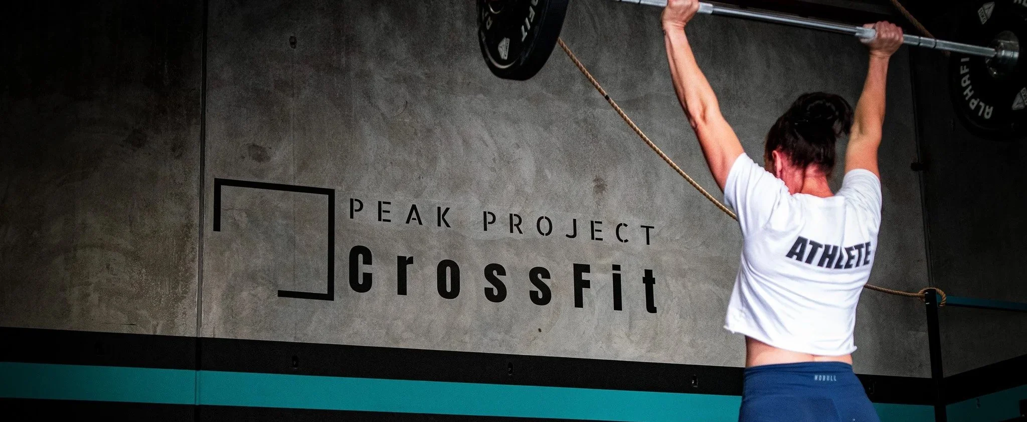 A woman lifting a barbell with weights at a CrossFit gym. She is wearing a white T-shirt with 'ATHLETE' on the back and blue shorts. The wall behind her has a sign that reads 'PEAK PROJECT CrossFit'.