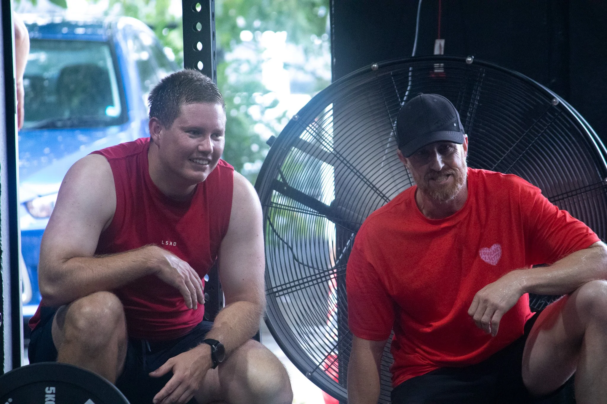 Two men wearing red shirts crouching next to a large electric fan during outdoor exercise.