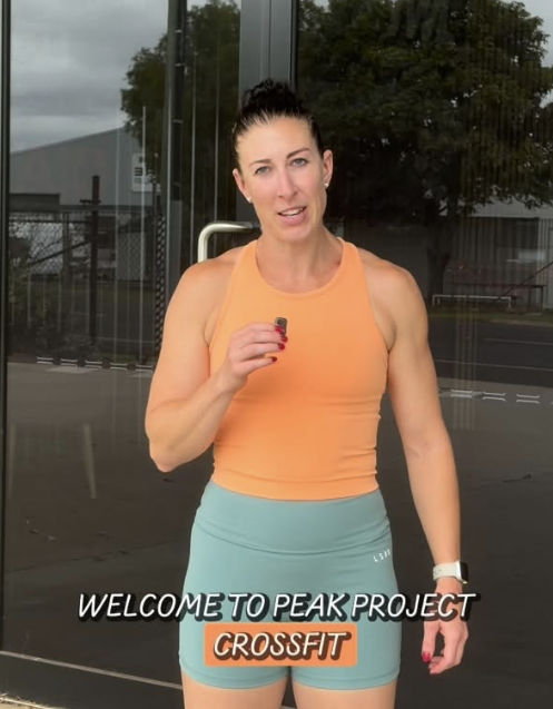 A woman in a peach crop top and blue shorts holding a small object, standing outside near a glass building with trees in the background, with text overlay 'WELCOME TO PEAK PROJECT CROSSFIT'.