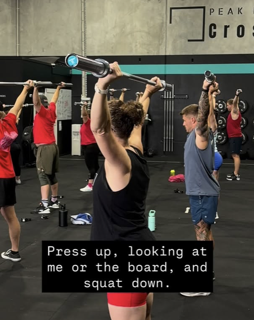 People participating in a group fitness class at a gym, lifting weights and doing squats, with an instructor demonstrating proper form.