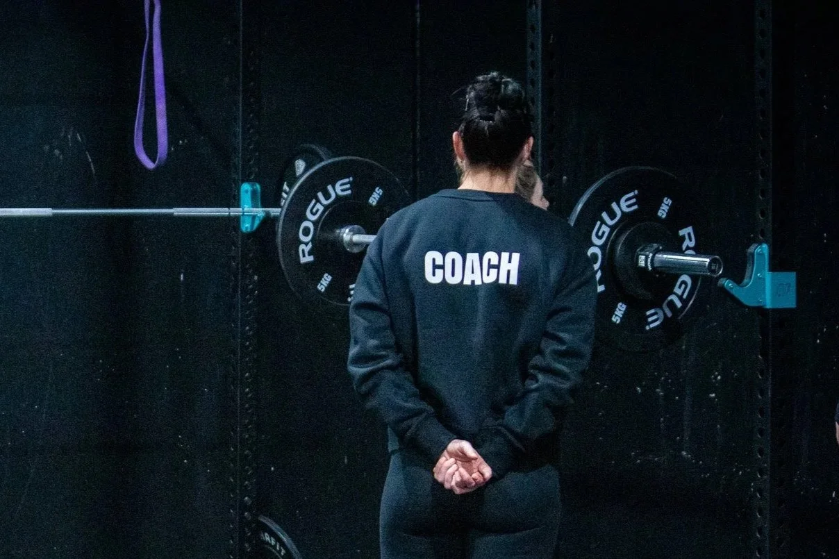 A woman with dark hair tied in a bun, wearing a black jacket with the word "COACH" written on the back, stands in front of a weightlifting barbell on a dark gym wall.