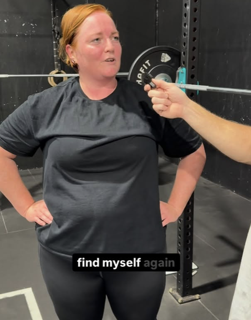 A woman with red hair and a black workout shirt standing with her hands on her hips, being interviewed or spoken to in a gym setting with weightlifting equipment in the background.