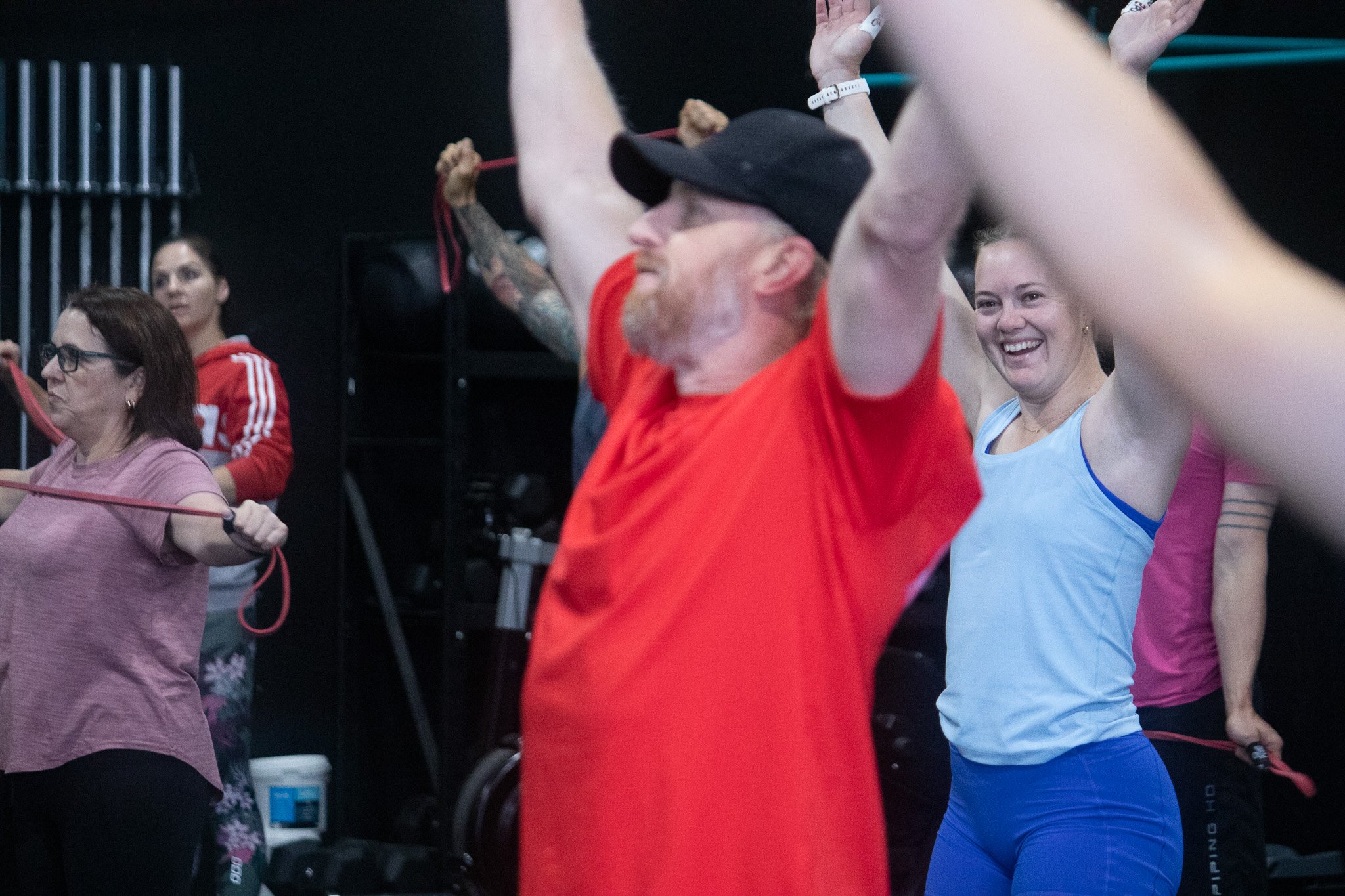 People exercising with resistance bands in a gym, including a woman smiling and a man with tattoos wearing a cap and orange shirt.