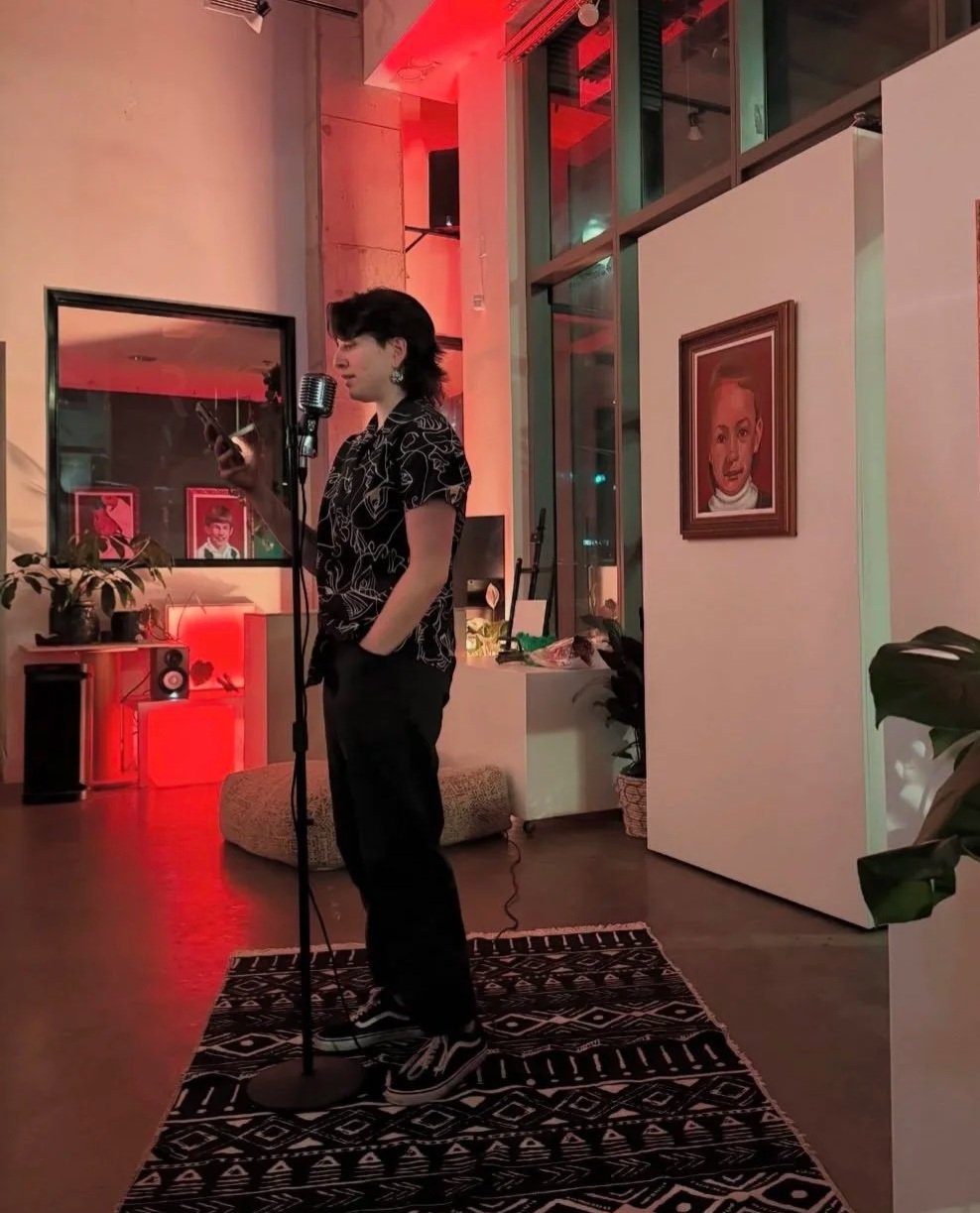 A person standing on a patterned rug, speaking or reading into a microphone, holding a phone, in a cozy, dimly lit room with warm red and orange lighting, artwork, plants, and a window.