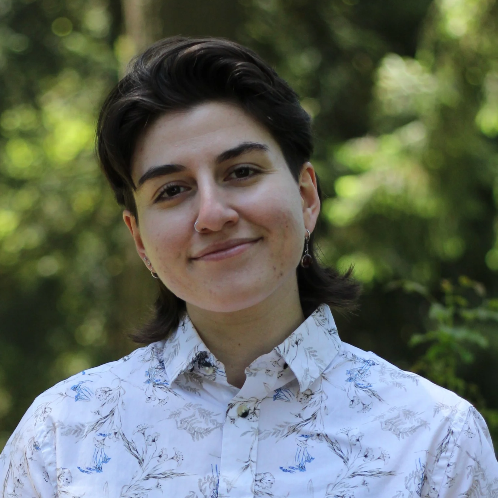 Young person with short dark hair, wearing a white shirt with a blue and gray floral pattern, standing outdoors with blurred greenery in the background.