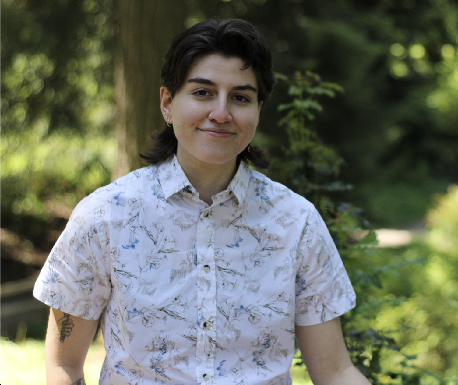 A young person with short dark hair, wearing a white button-up shirt with a floral pattern, smiling outdoors in a green, wooded area.