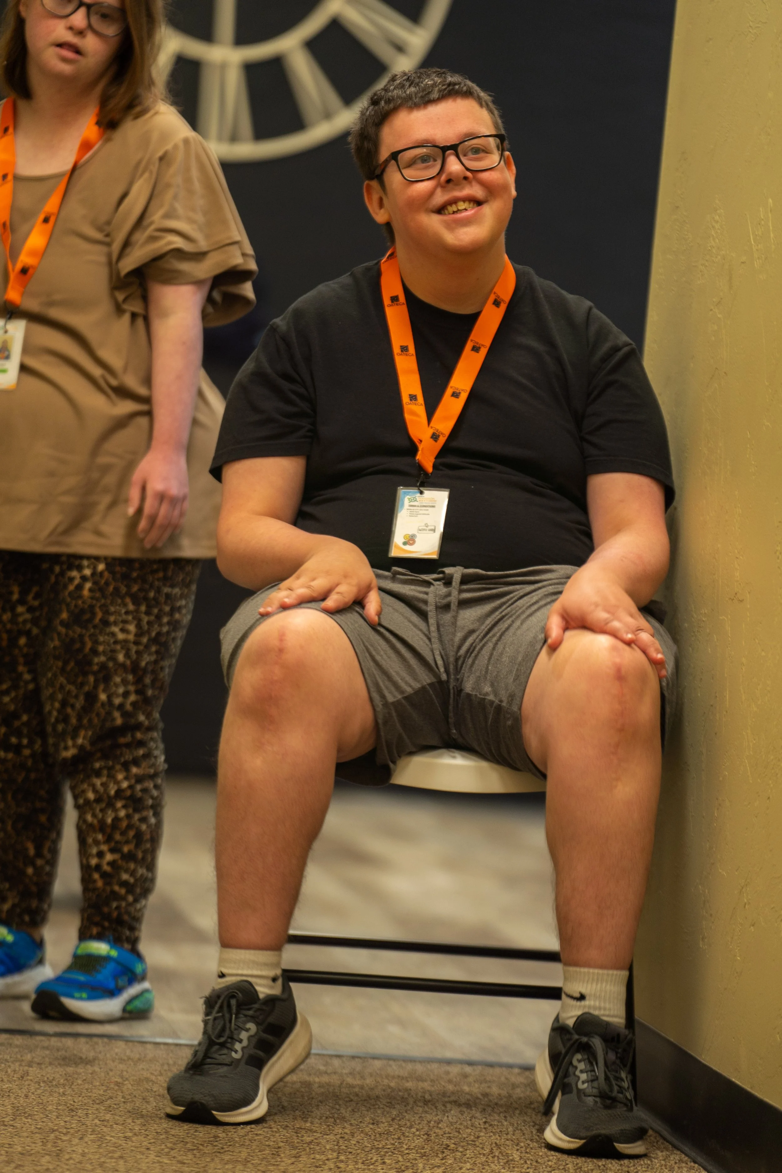 A young man with glasses sitting on a toilet, smiling, with skin condition scars on his legs. He wears a black t-shirt, gray shorts, white socks, and black sneakers. There is a woman standing beside him, wearing leopard print pants and a tan top. The setting appears to be a public or institutional bathroom with a yellow wall and a clock in the background.