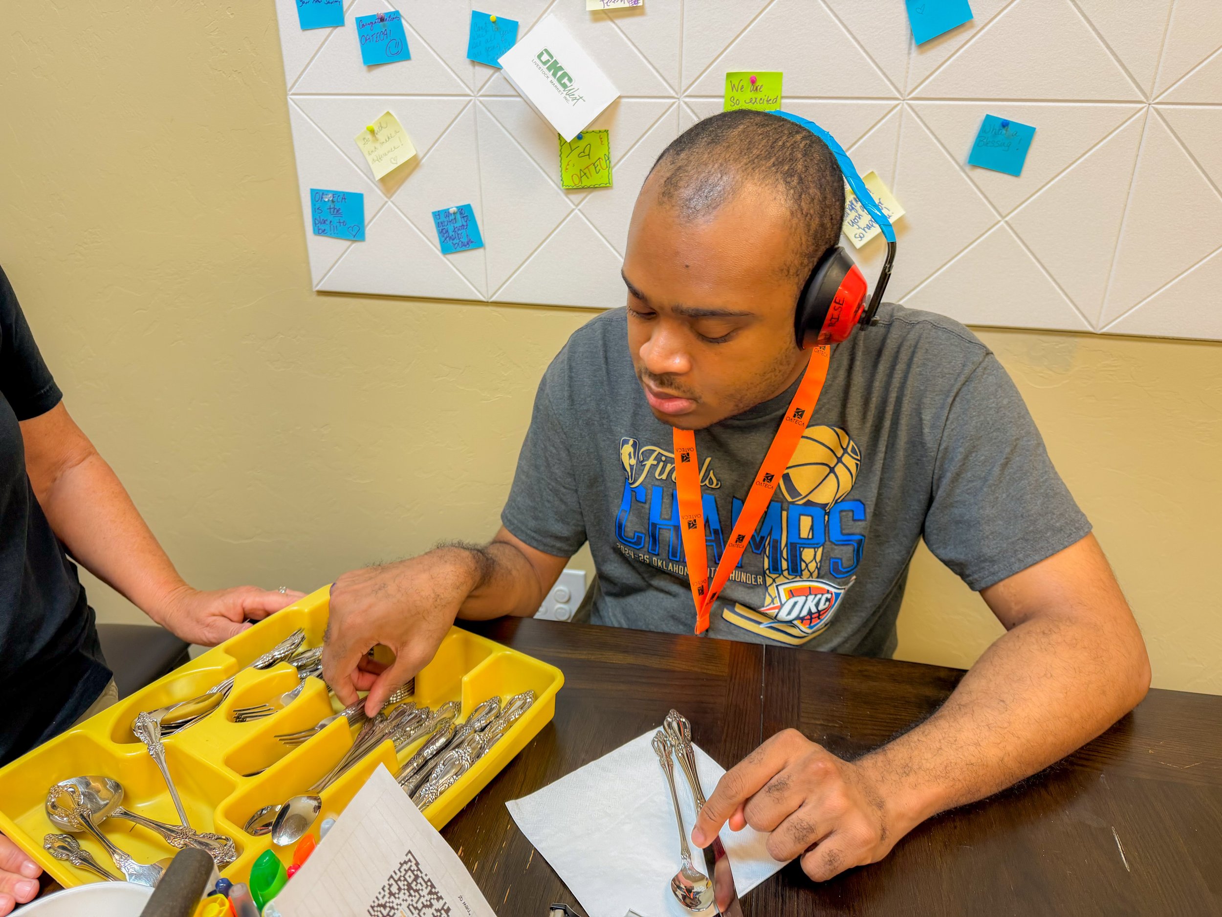 A young man wearing a Oklahoma City Thunder basketball T-shirt and red headphones is sitting at a table, looking down. There are yellow containers with silverware on the table, and his hand is reaching into one container. In the background, there is a white tile wall with various colorful sticky notes and papers attached.