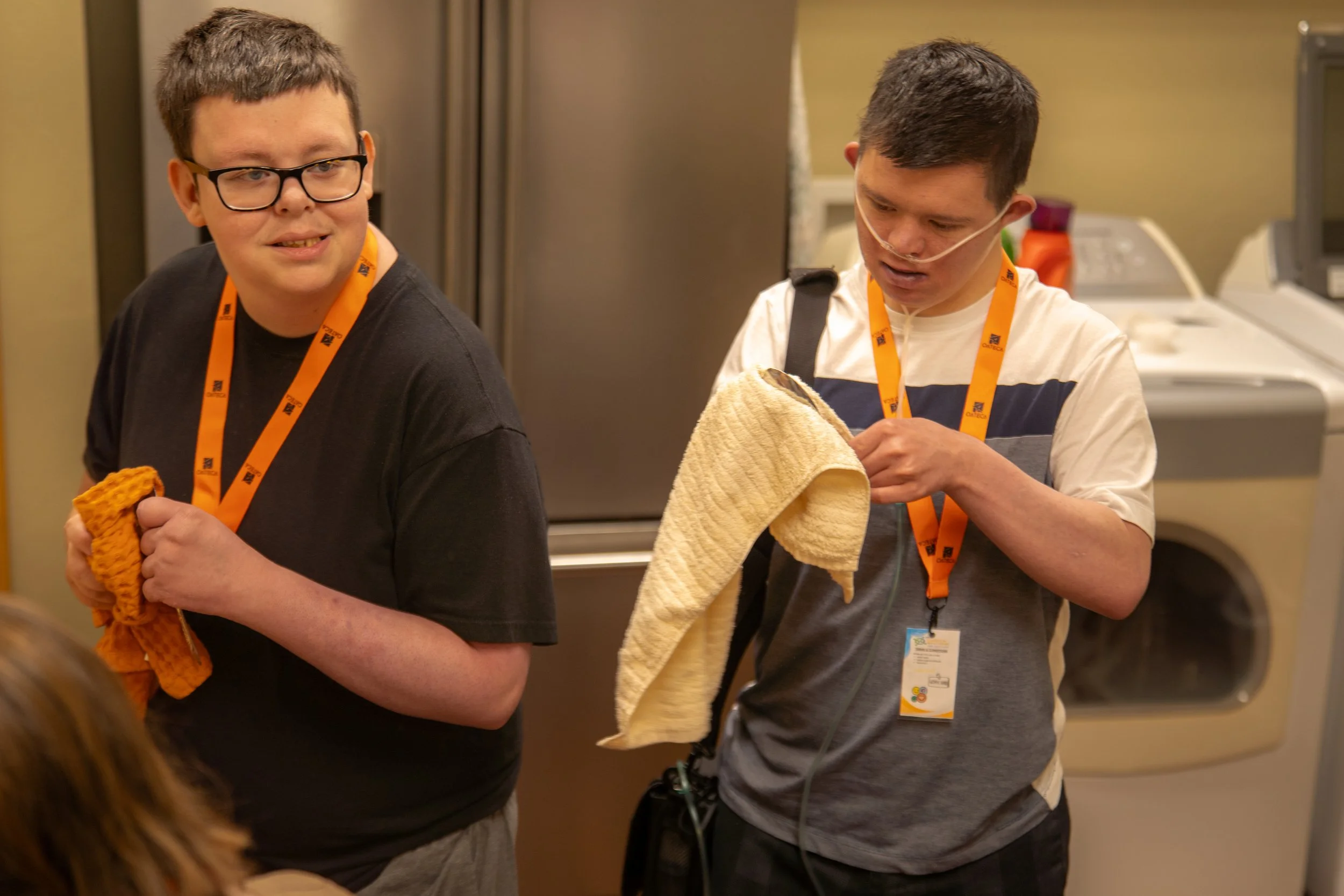 Two young men in a kitchen, one wearing glasses and holding an orange cloth, and the other with an oxygen tube, holding a beige towel. Both are wearing orange lanyards with a logo.