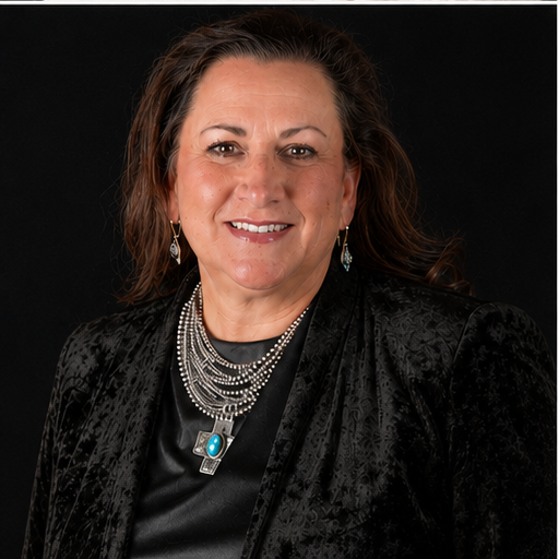 A woman with shoulder-length dark hair, wearing a black textured blazer, a black top with a silver necklace, and turquoise jewelry, smiling against a black background.