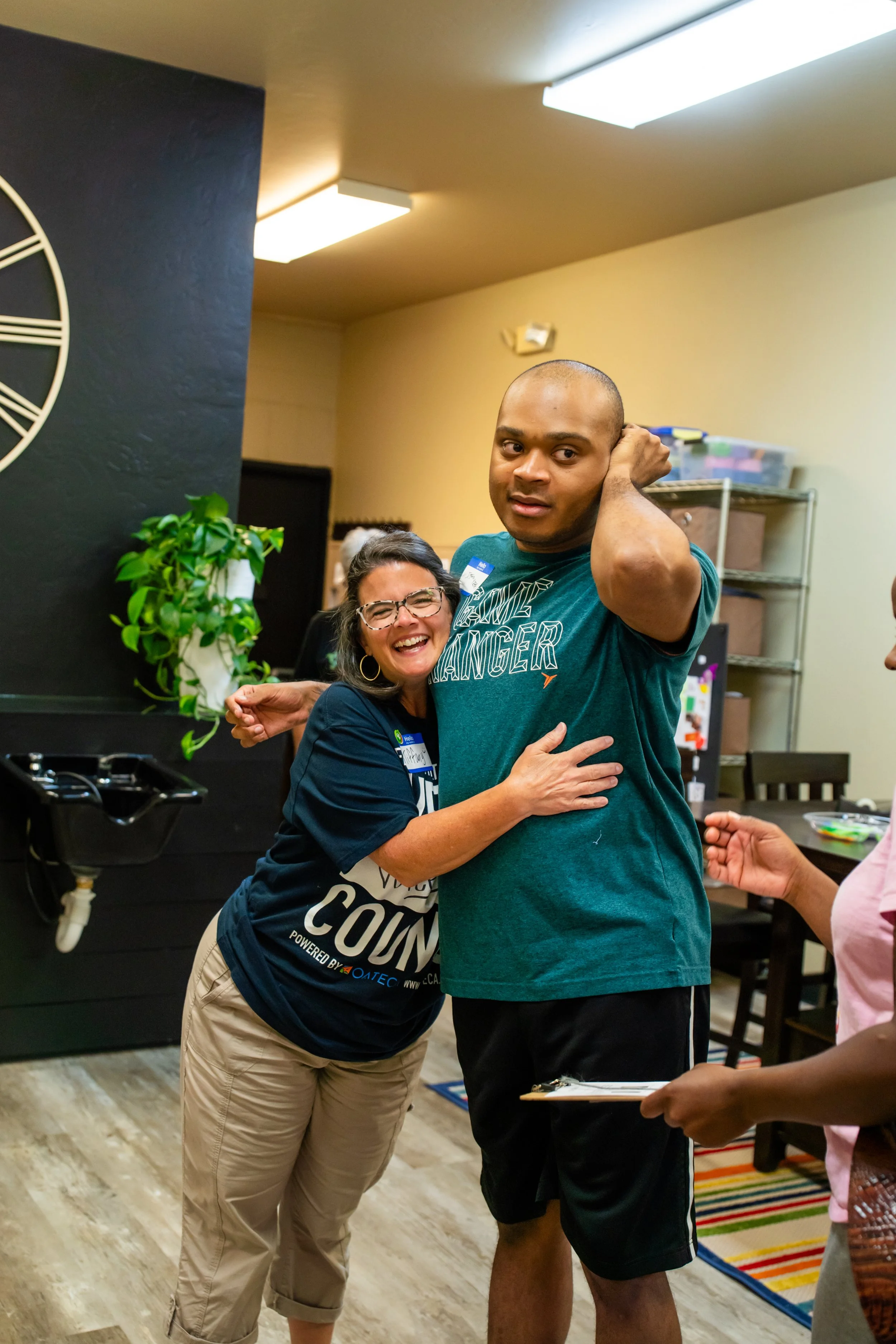 A woman and a man hugging and smiling at an indoor gathering. The woman is wearing glasses and a blue t-shirt, while the man is in a teal t-shirt and black shorts. They are in a room with fluorescent lighting, a black wall with a clock, and shelves with storage boxes in the background.