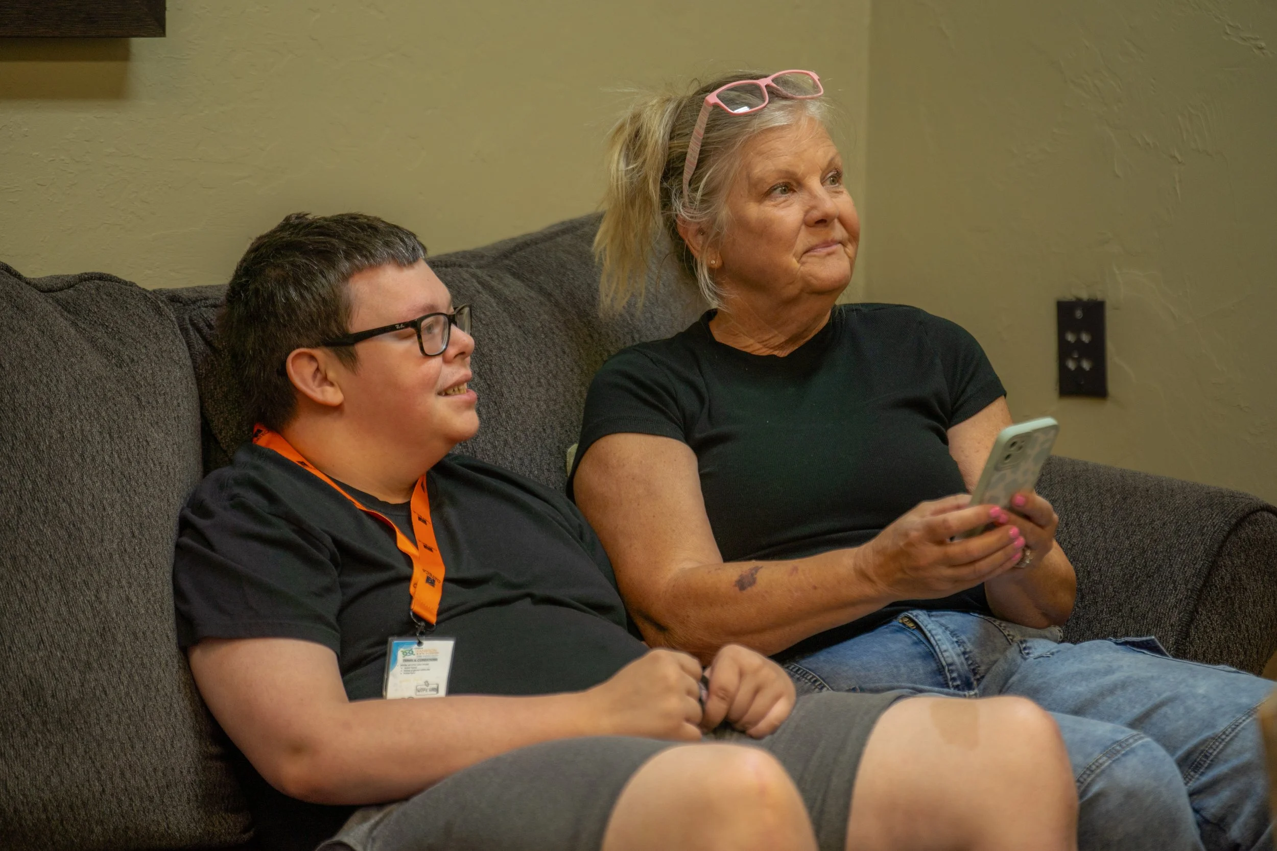A woman with grey hair and glasses on her head sitting on a couch, holding a smartphone and looking attentively. Beside her, a young man with glasses in a black shirt and an orange lanyard, also sitting on the couch, smiling and looking in the same direction.