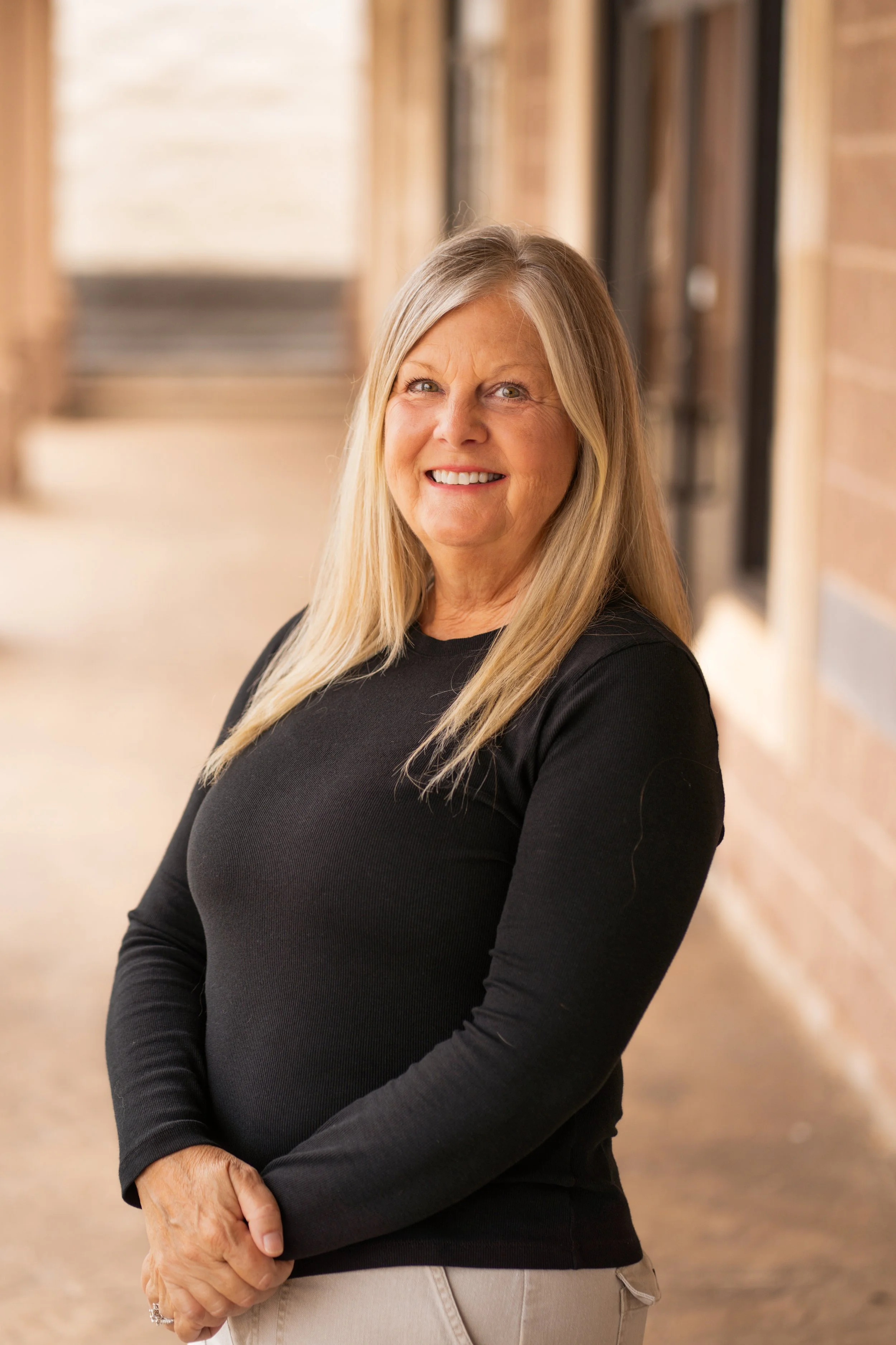 A smiling woman with long gray hair wearing a black long sleeve top standing in a hallway with beige walls and stairs in the background.