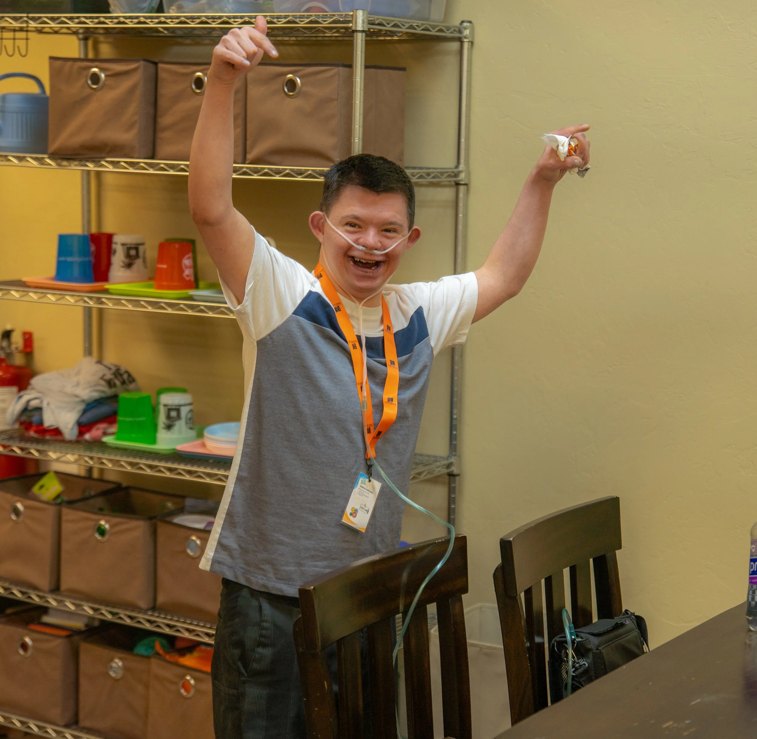 A person with a nasal cannula smiling and raising one arm, standing in a room with metal shelving and containers in the background.