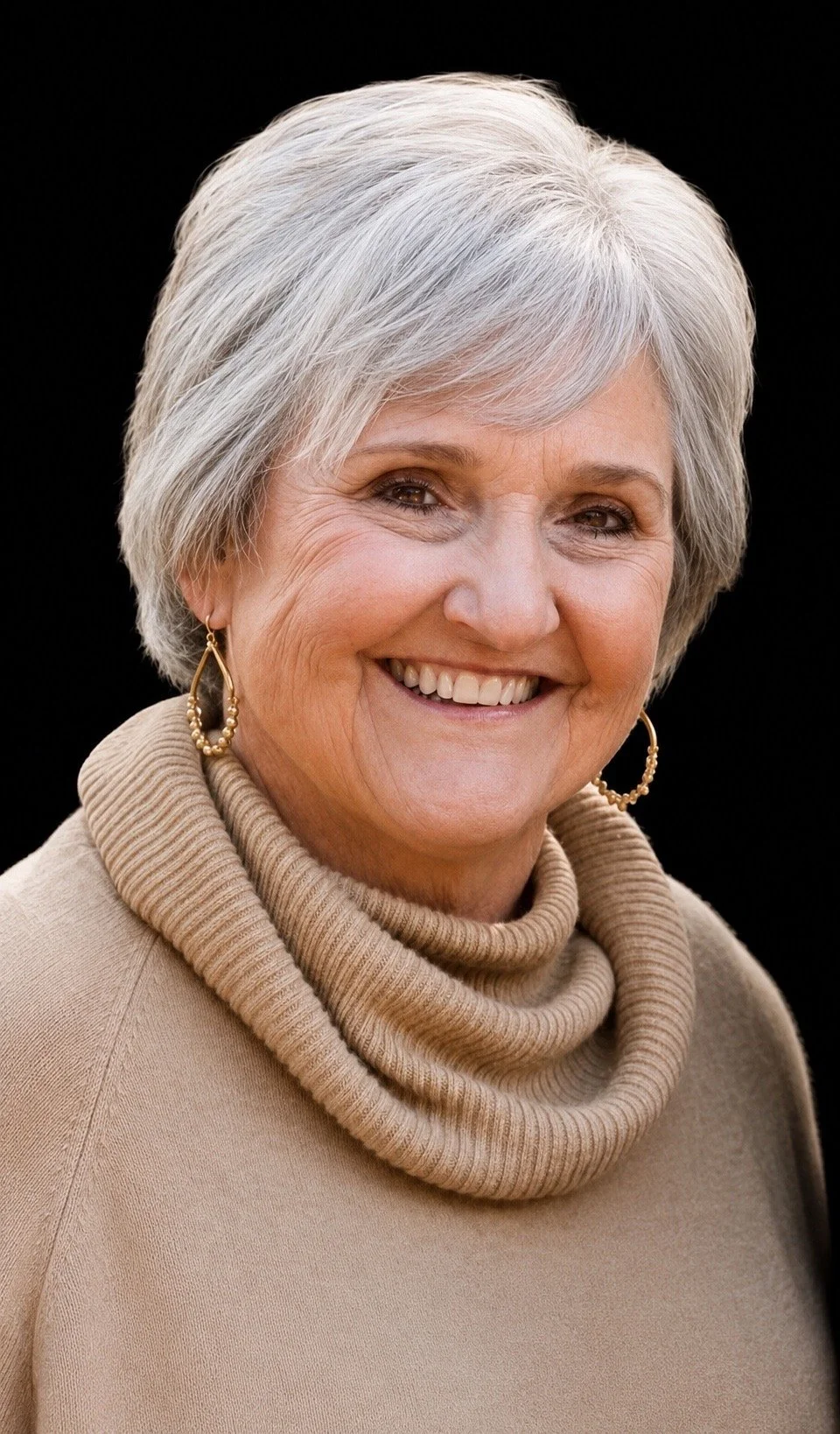 A smiling elderly woman with short gray hair, wearing a beige turtleneck sweater and gold hoop earrings, against a black background.