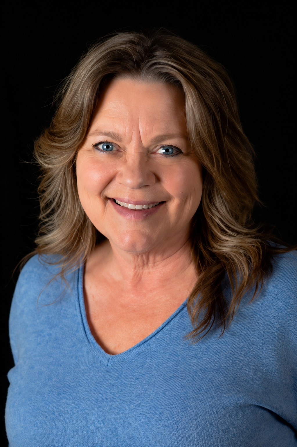 Portrait of a smiling middle-aged woman with blue eyes, shoulder-length wavy brown hair, wearing a light blue V-neck shirt, against a black background.