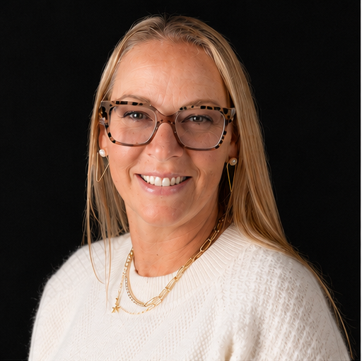 Portrait of a woman with long blonde hair, wearing tortoiseshell glasses, a white sweater, and gold jewelry, smiling against a black background.