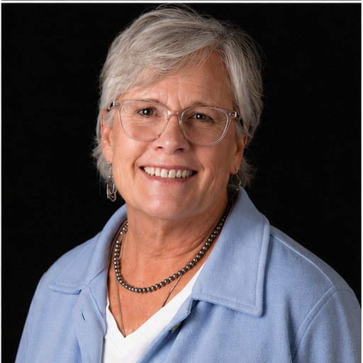 A portrait of an older woman with short gray hair, wearing glasses, a light blue jacket, a white shirt, and a necklace, smiling against a black background.