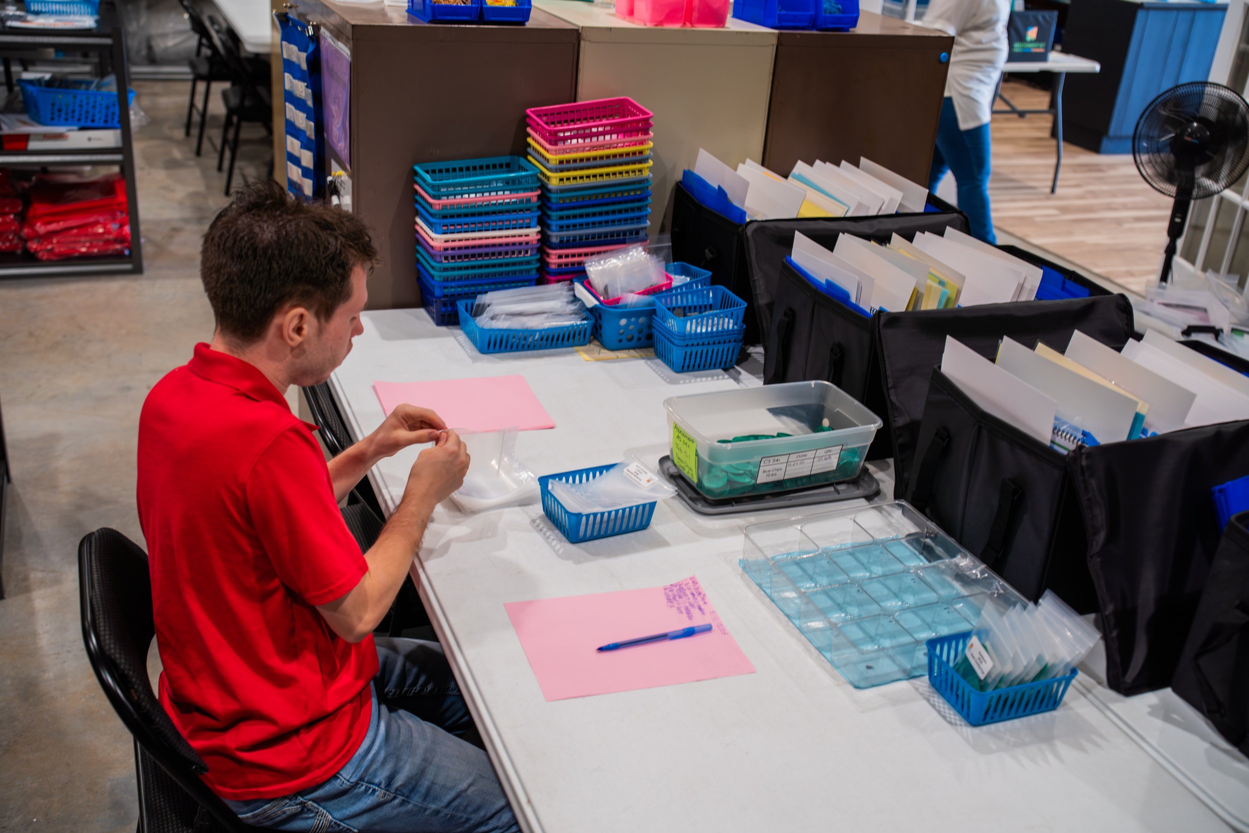 A man in a red shirt sitting at a table with organized containers and folders, working at an office or workspace environment.