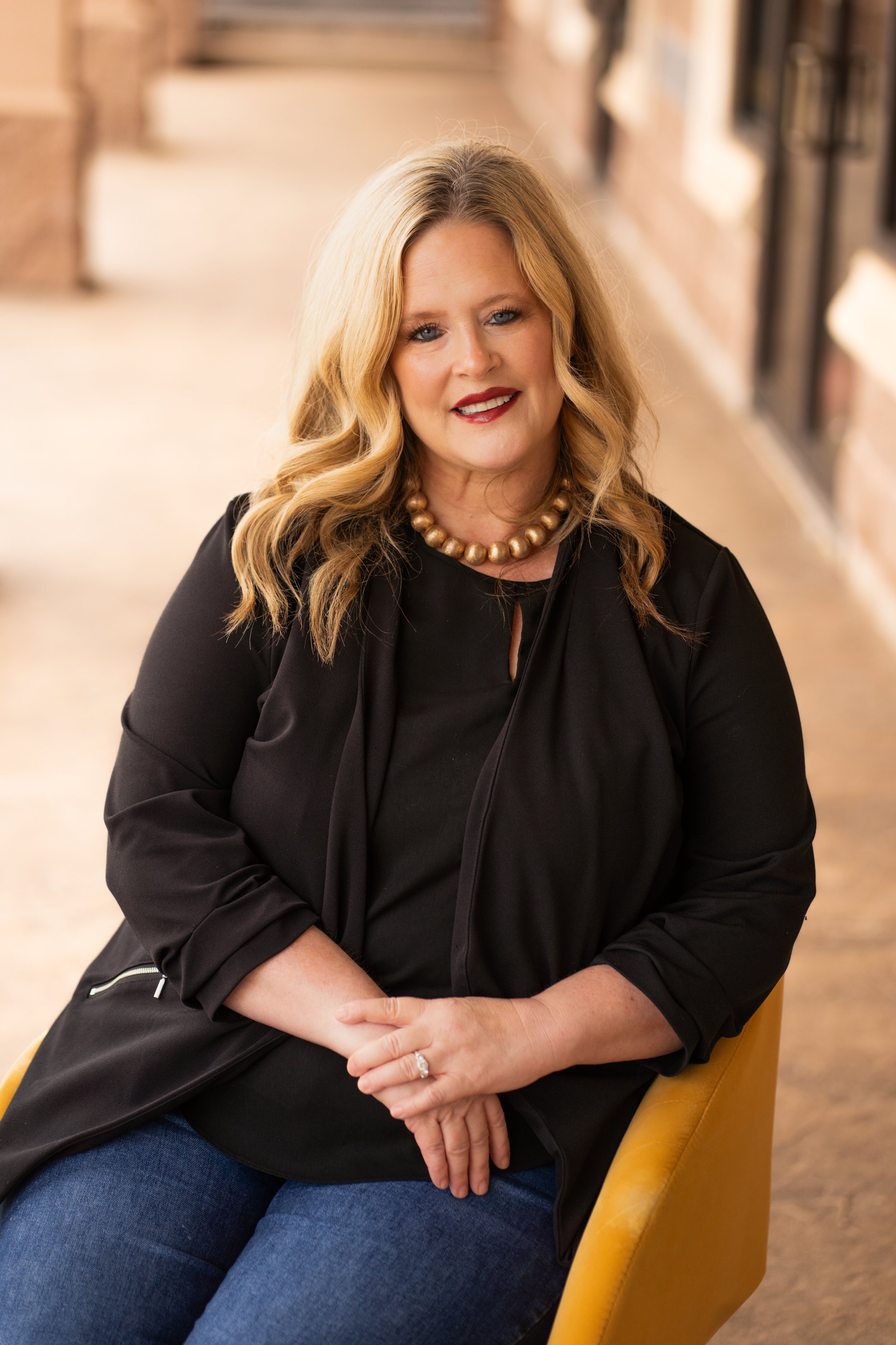 A woman with blonde, wavy hair and blue eyes wearing a black blazer and a gold necklace, sitting on a yellow chair in a cozy indoor space with a brick wall and bookshelves in the background.