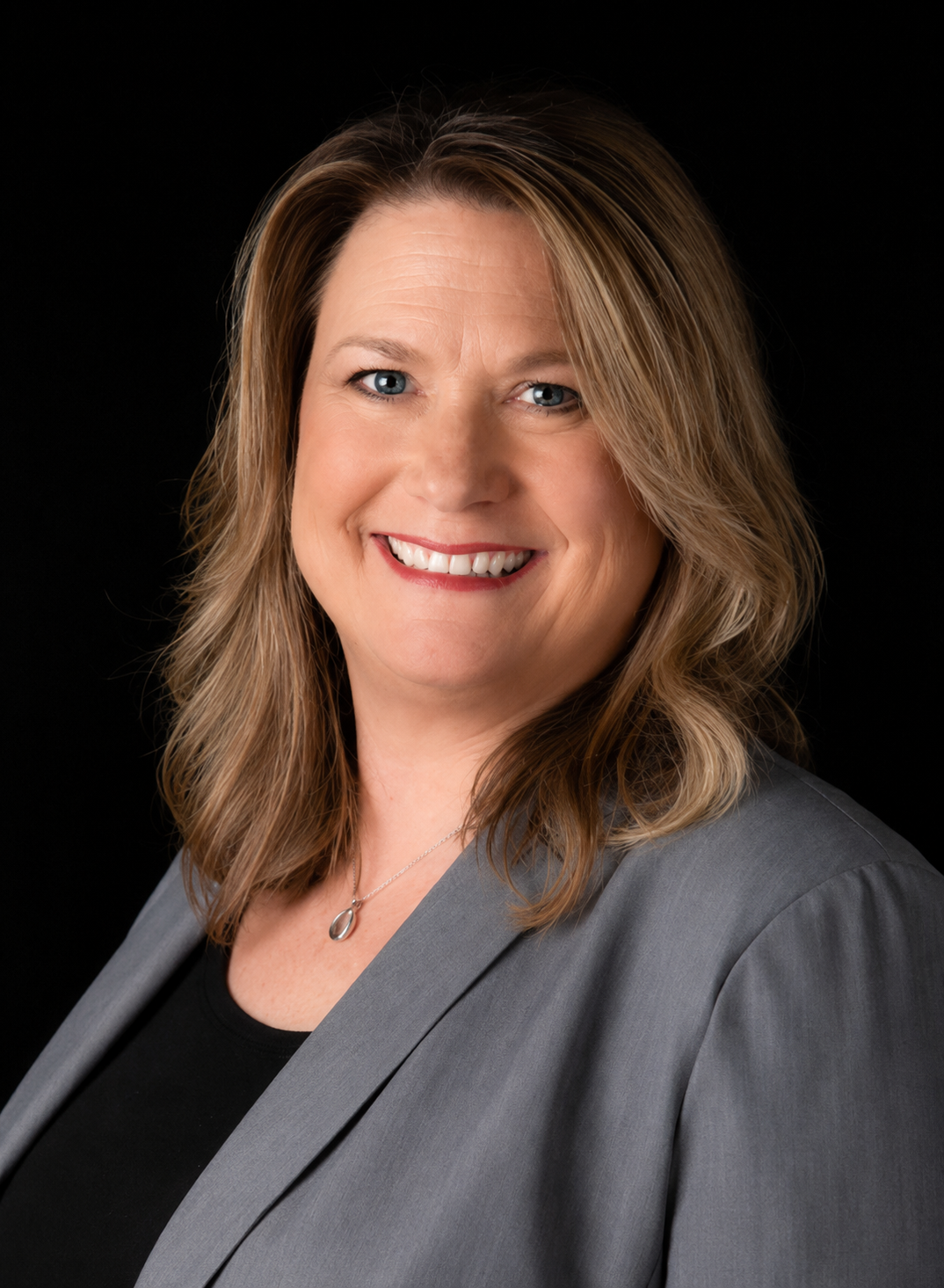 Headshot of a woman with shoulder-length wavy blonde hair, wearing a gray blazer over a black top, smiling against a black background.