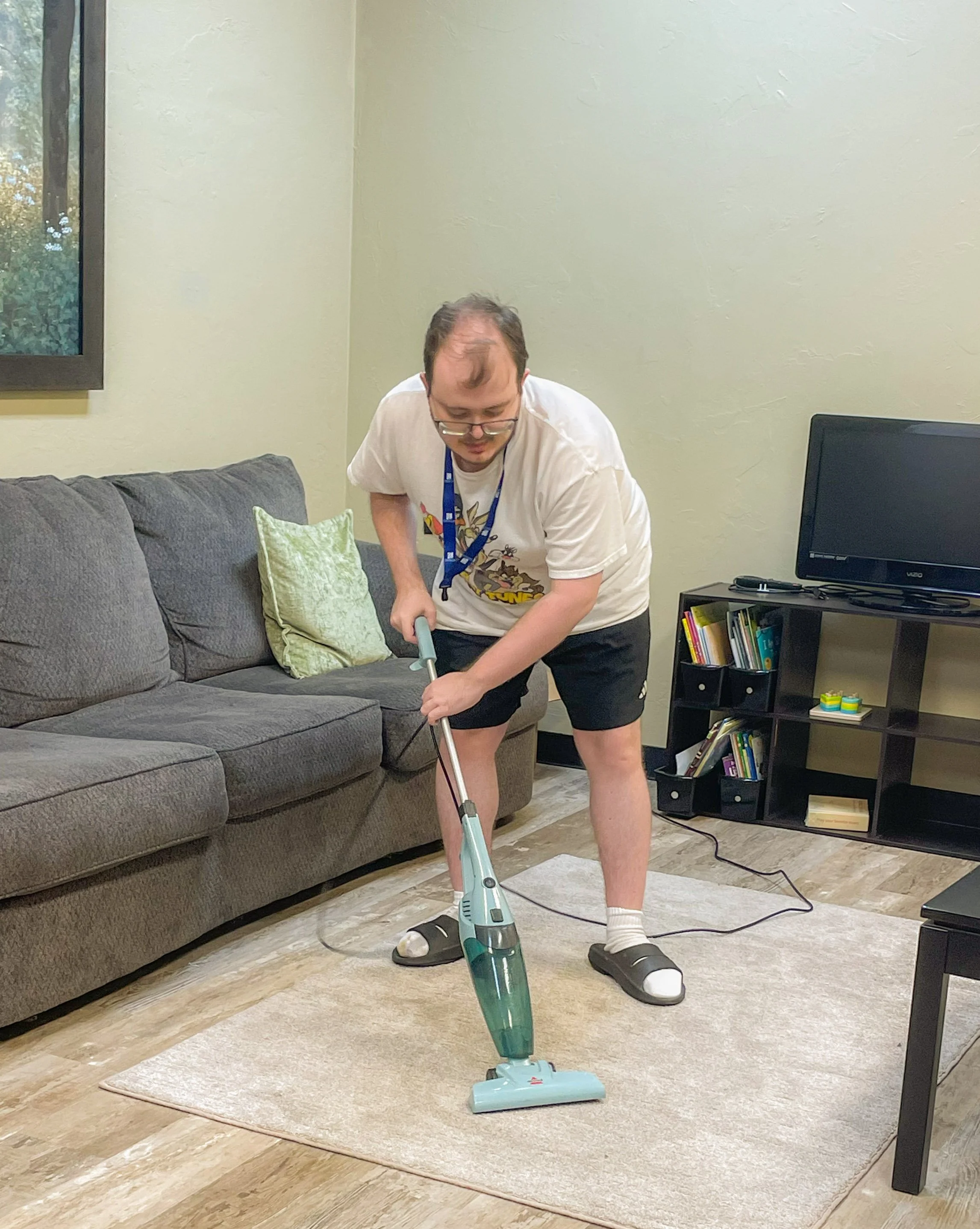 A man is vacuuming a rug in a living room. The room has a gray sofa with a green pillow, a window, a black bookshelf filled with books, and a wall with a light color. The man is wearing a white T-shirt with cartoon characters, shorts, and socks with slippers.