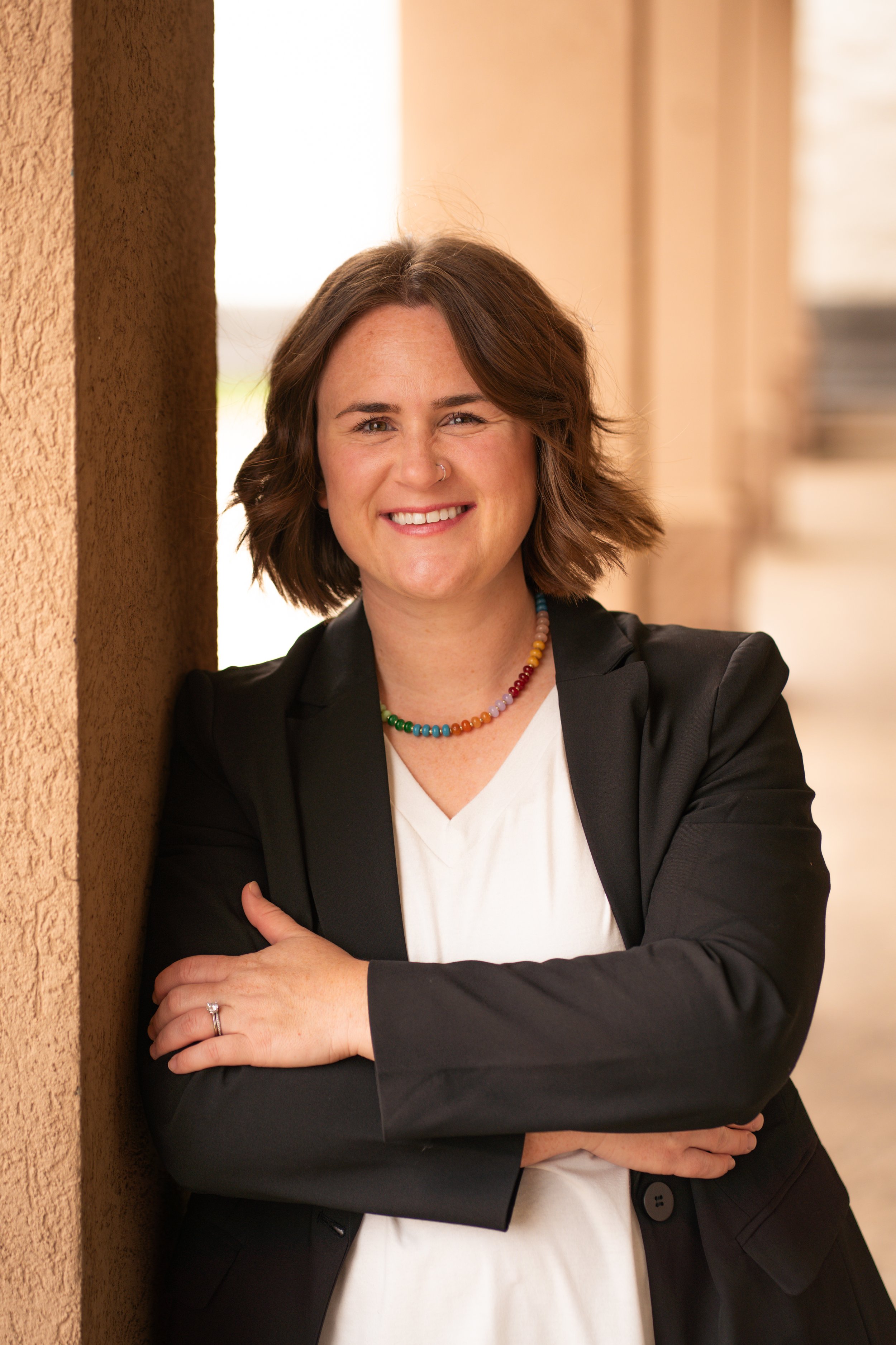 A woman with short brown hair smiling, wearing a black blazer, white shirt, and a colorful beaded necklace, standing with her arms crossed, leaning against a textured wall in an indoor setting.