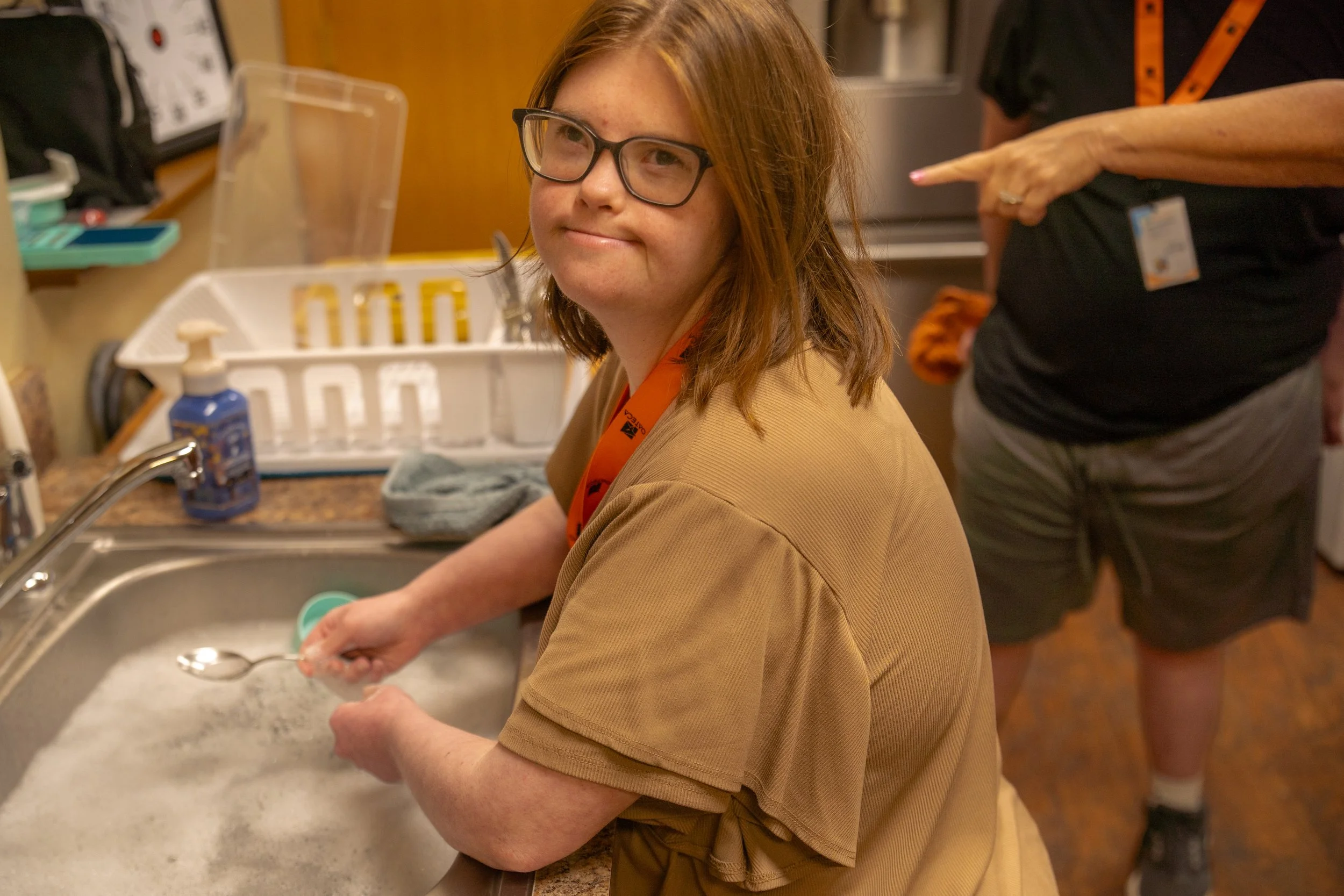 A young girl with red hair, glasses, and a tan shirt is washing dishes at a kitchen sink. An adult woman, wearing a black shirt and a name badge, stands nearby. The kitchen has a white dish rack, soap on the counter, and various kitchen items.