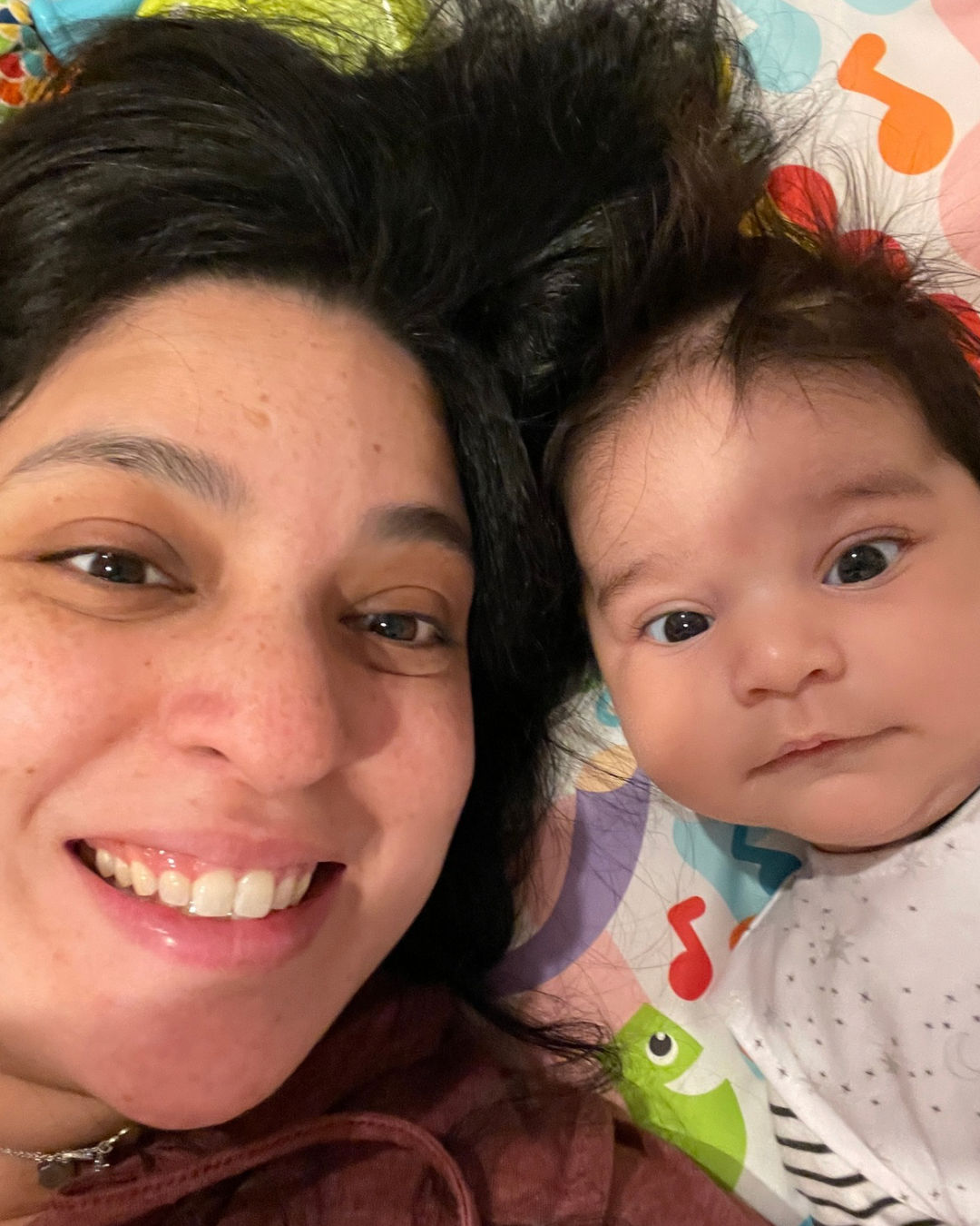 A woman and a baby lying down with their heads close together, smiling and looking at the camera. The woman has dark hair and the baby has dark hair with some curls, lying on a colorful mat with musical notes and a frog design.