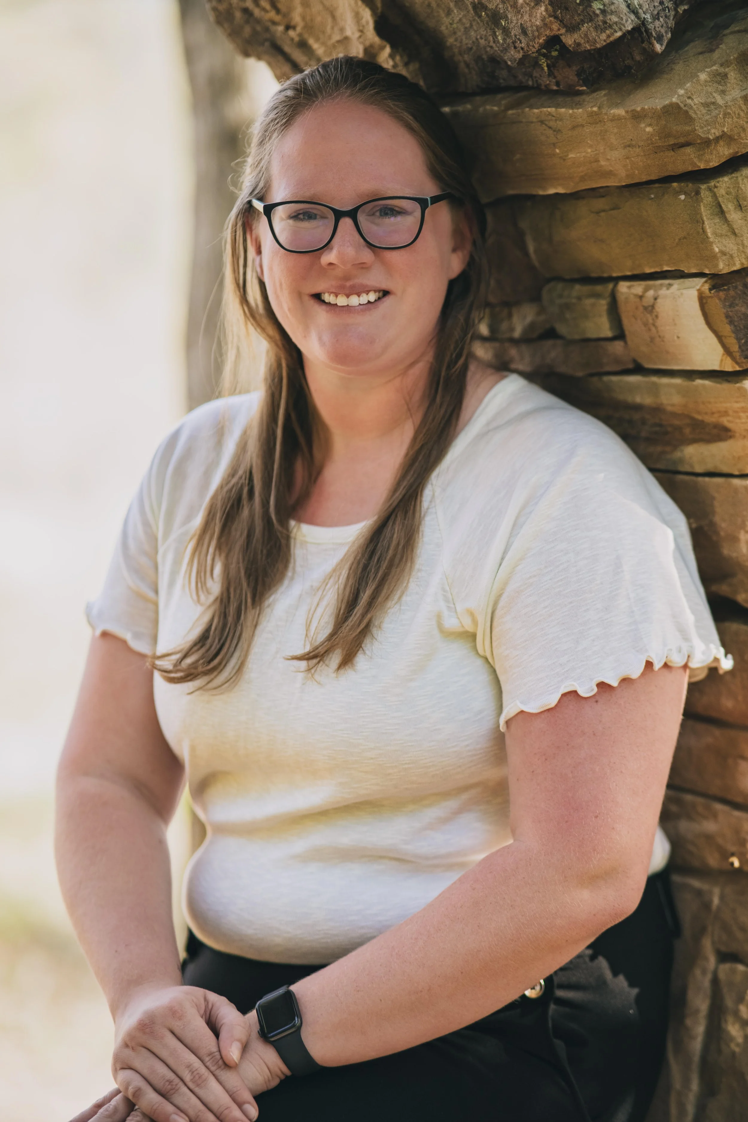 A woman with long hair, wearing glasses and a white t-shirt, smiling and leaning against a stone wall outdoors.