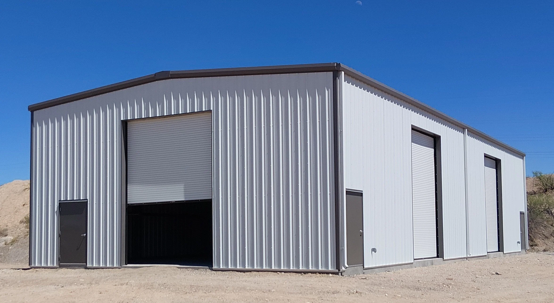 A modern metal storage building with two roll-up doors on a dirt lot under a clear blue sky.