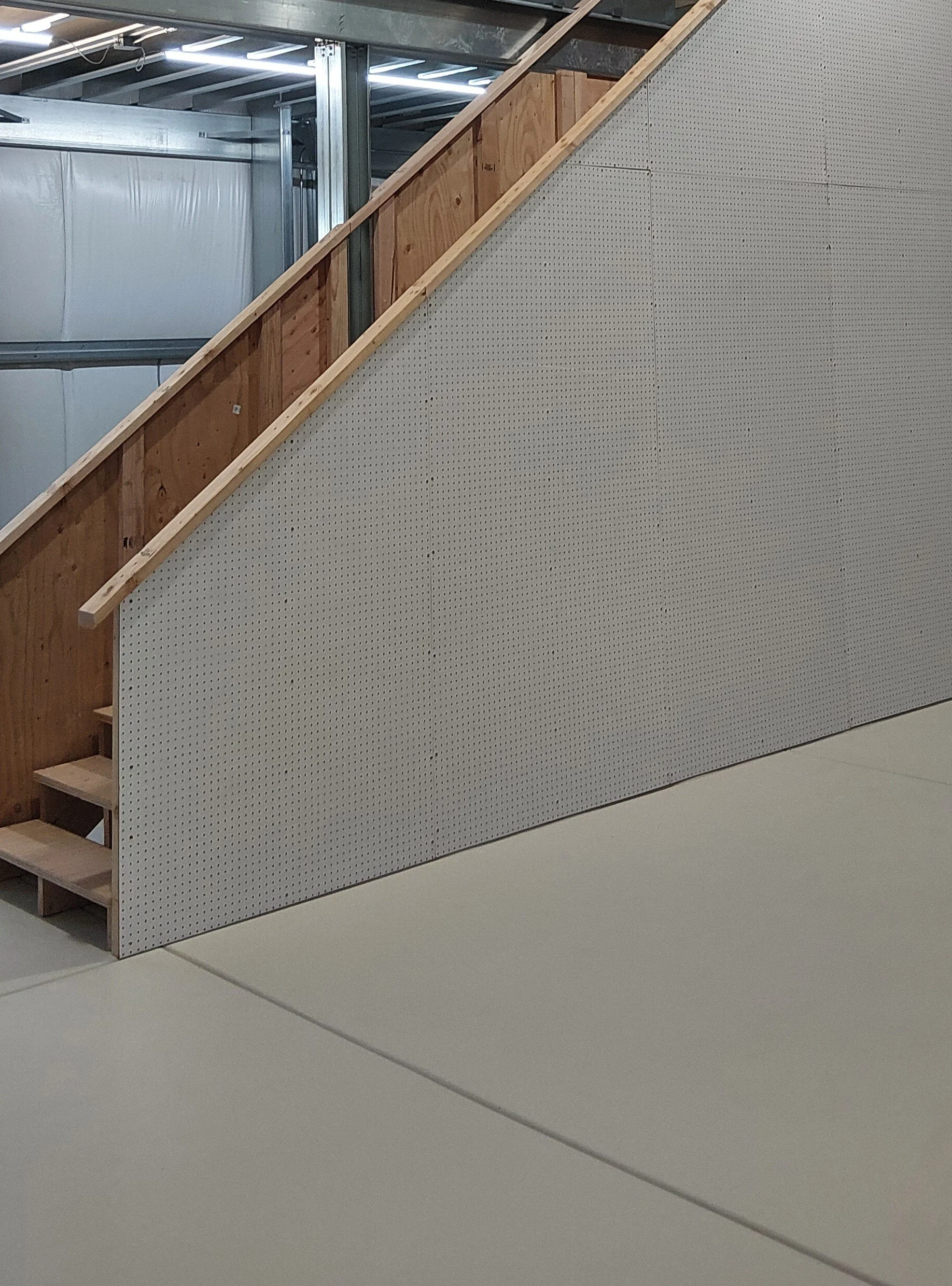 Interior of a building under construction with a partially built stairway, unfinished wooden framing, and white pegboard wall panels.
