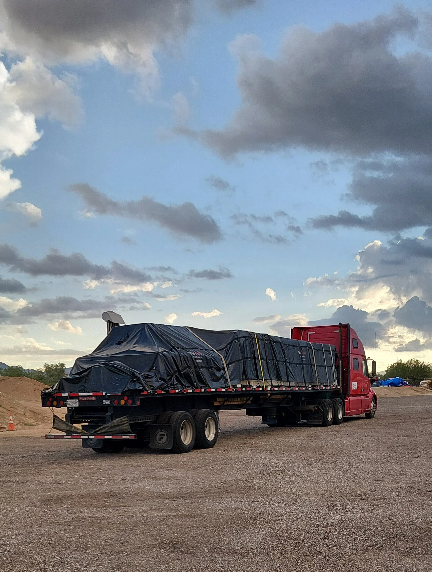 A flatbed truck carrying a steel load under black tarp, parked under a cloudy sky.