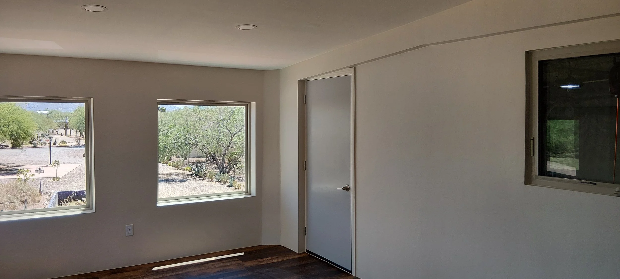 Interior of a room with two large windows showing a desert landscape with trees and a wall in the background, a white door, and a small window on the right, with dark wood flooring.