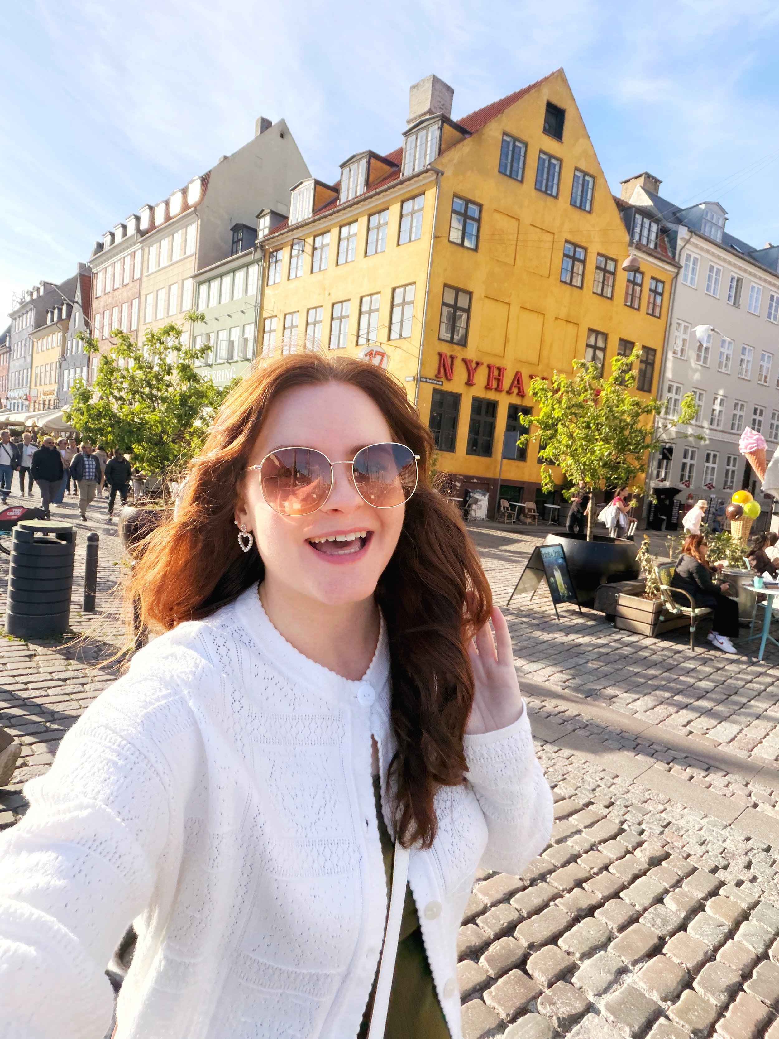A woman with long red hair wearing sunglasses and a white sweater, smiling, standing in a European square with colorful buildings in the background, and people walking around.