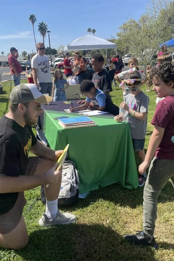 Children and adults at an outdoor event with a green table, where children are signing papers and receiving water bottles, under white canopy tents with palm trees in the background on a sunny day.