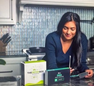Woman with long dark hair sitting at a kitchen counter, looking at a card, with a DNA testing kit and detox box in front of her.