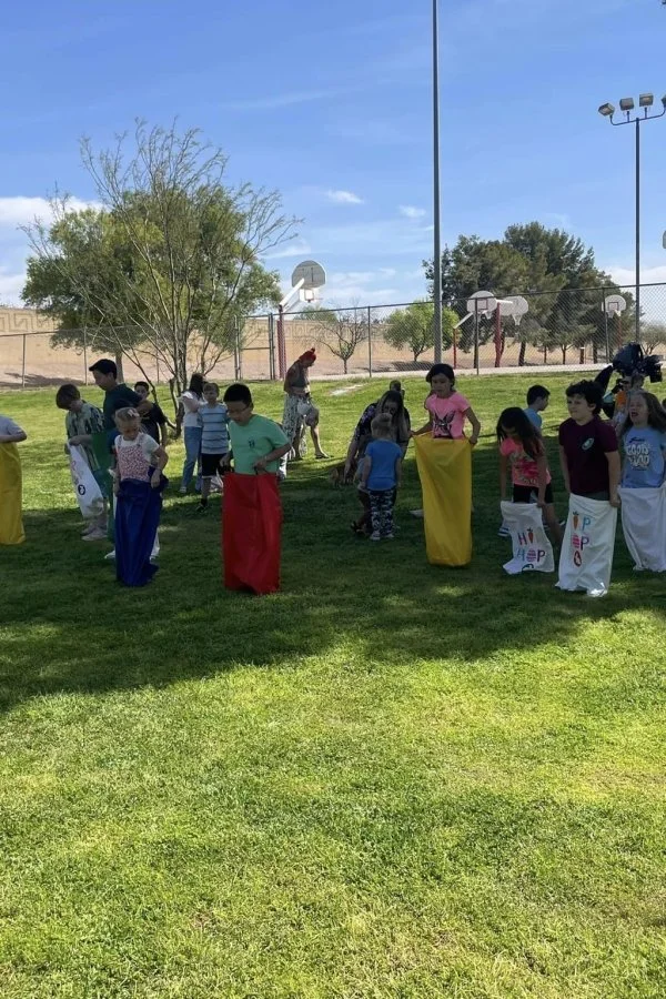 Children participating in a sack race outdoors, with some holding decorated sacks, on a grassy field near a playground with basketball hoops, trees, and a fence.